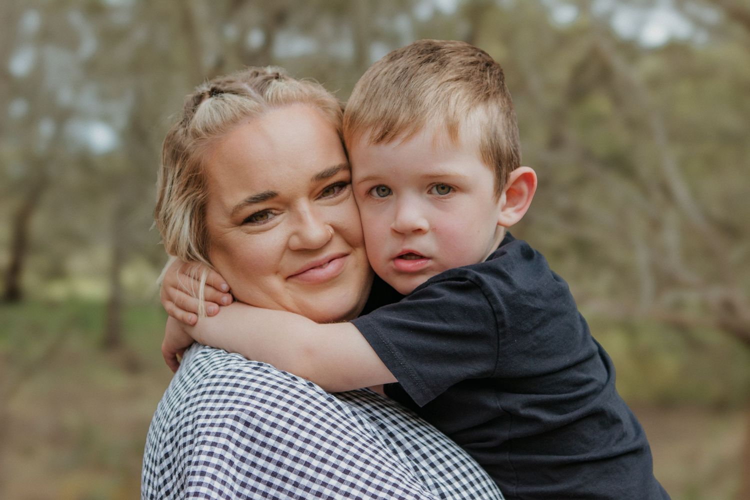 Woman smiling while a young boy hugs her from behind in a natural outdoor setting.