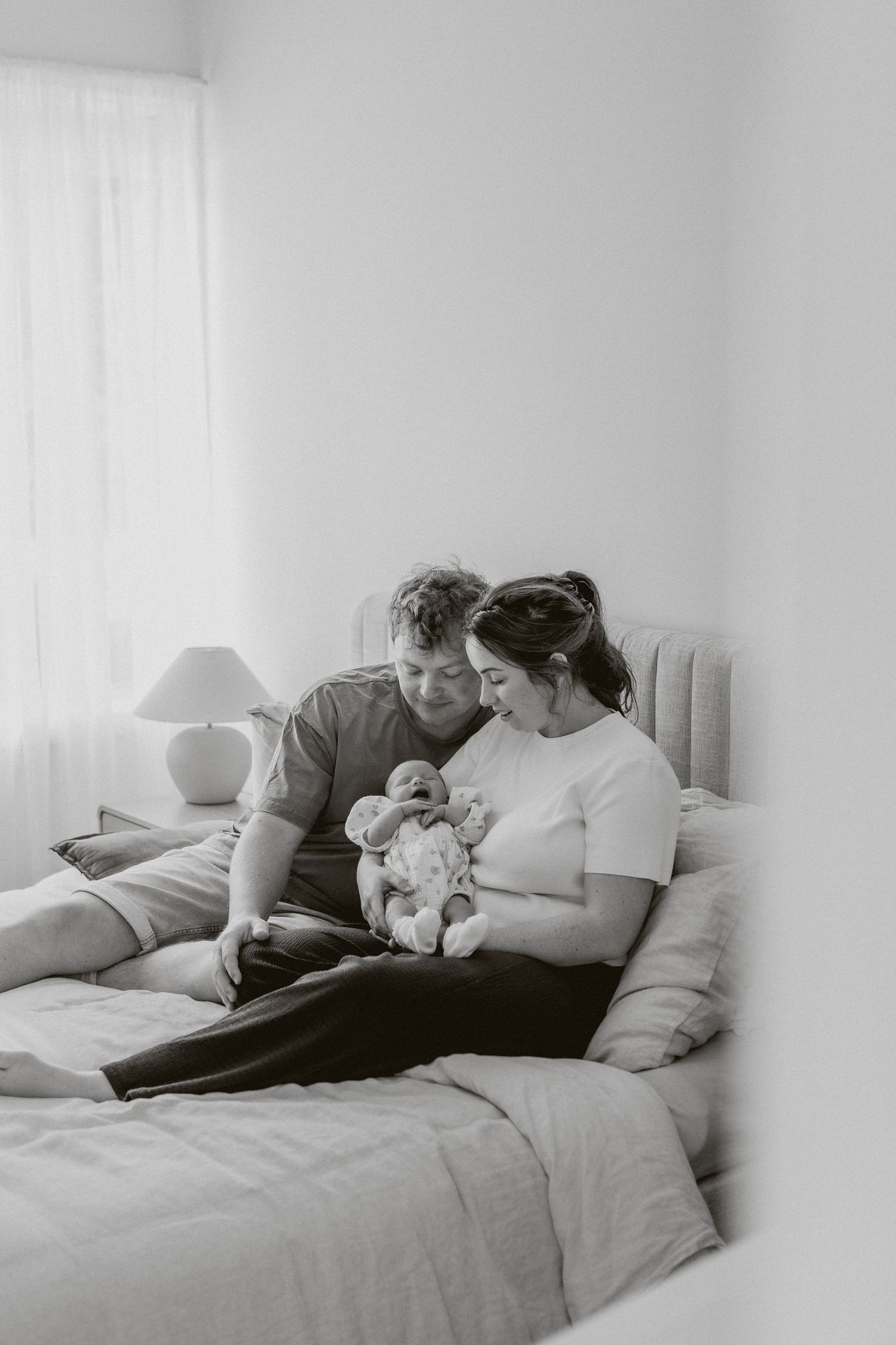 Black and white photo of new parents sitting on a bed together lovingly gazing at their newborn.