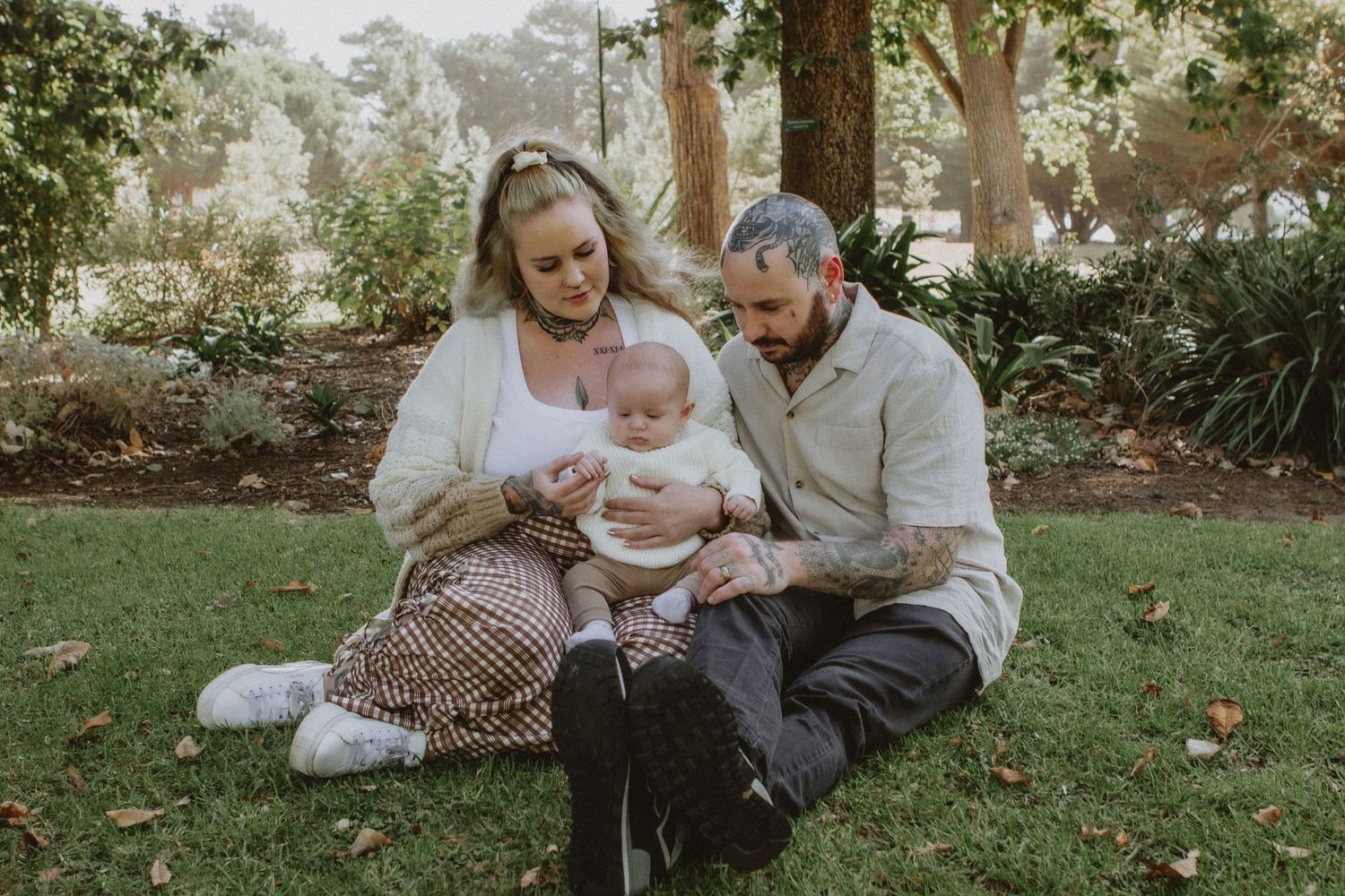 Young couple sitting on grass in park cradling newborn baby surrounded by autumn trees at Warrnambool Botanical Gardens.