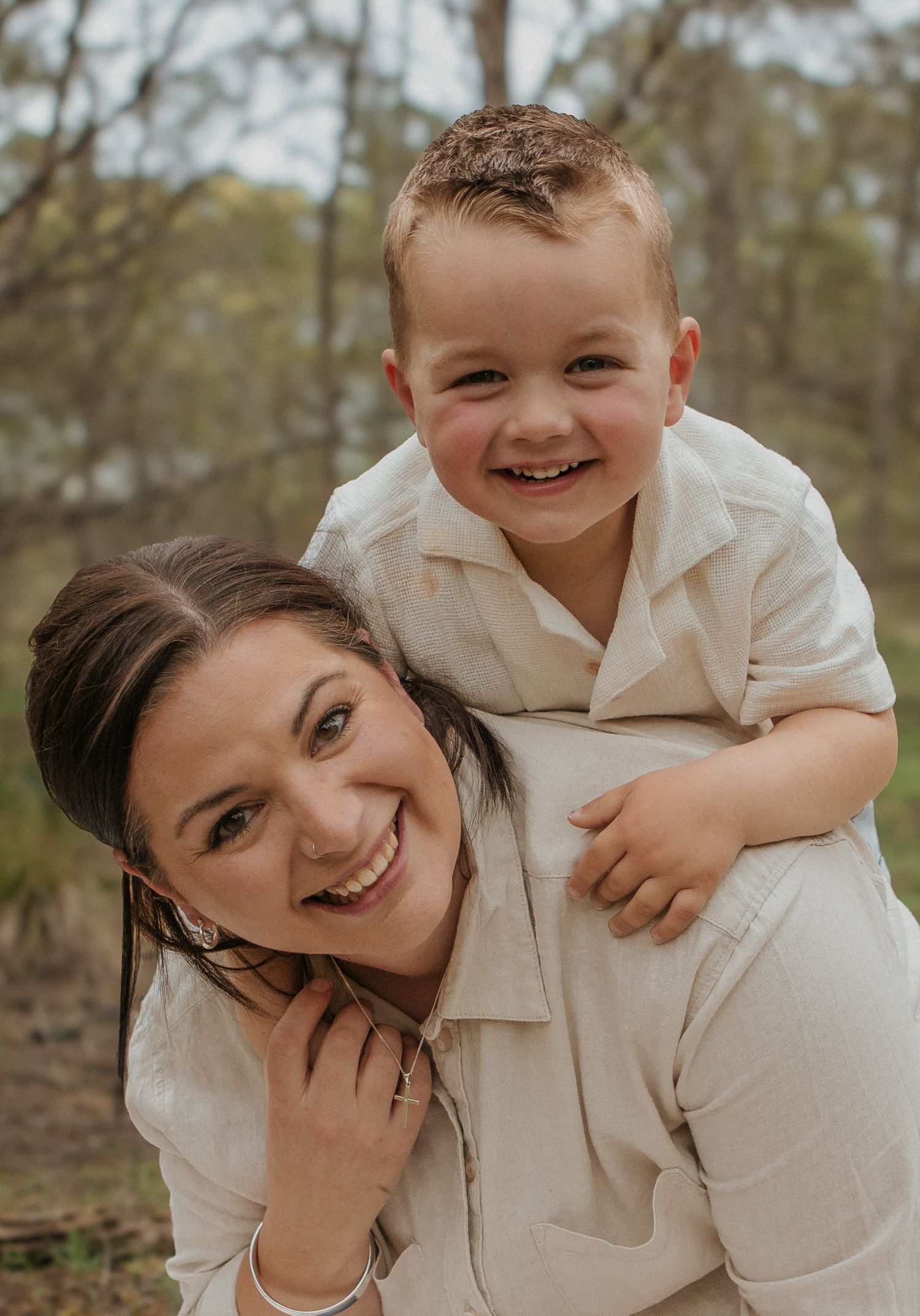 A smiling woman giving a happy toddler boy a piggyback ride outdoors in nature.