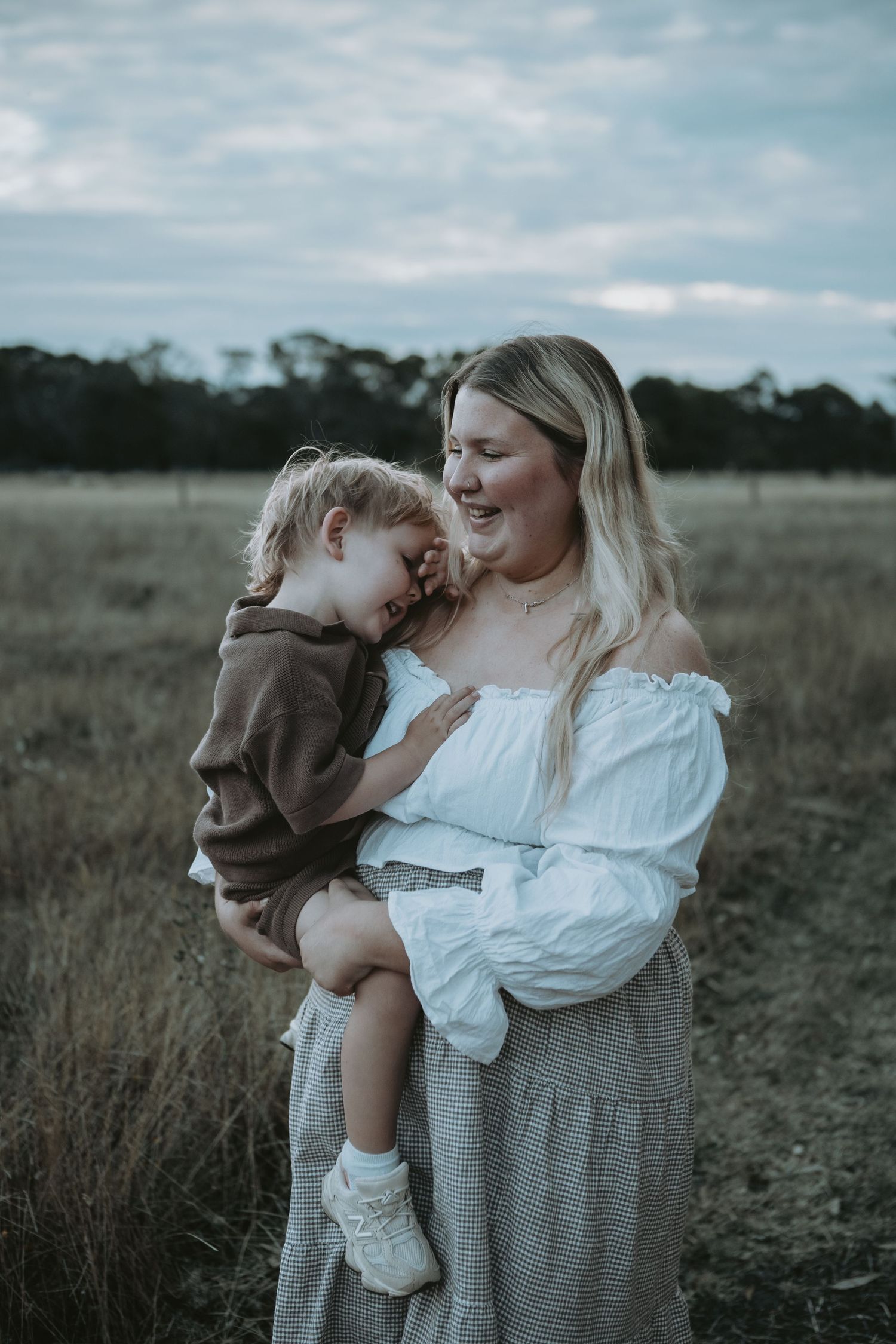 A mother in a white off-shoulder top holds her laughing toddler in a dry grass field.