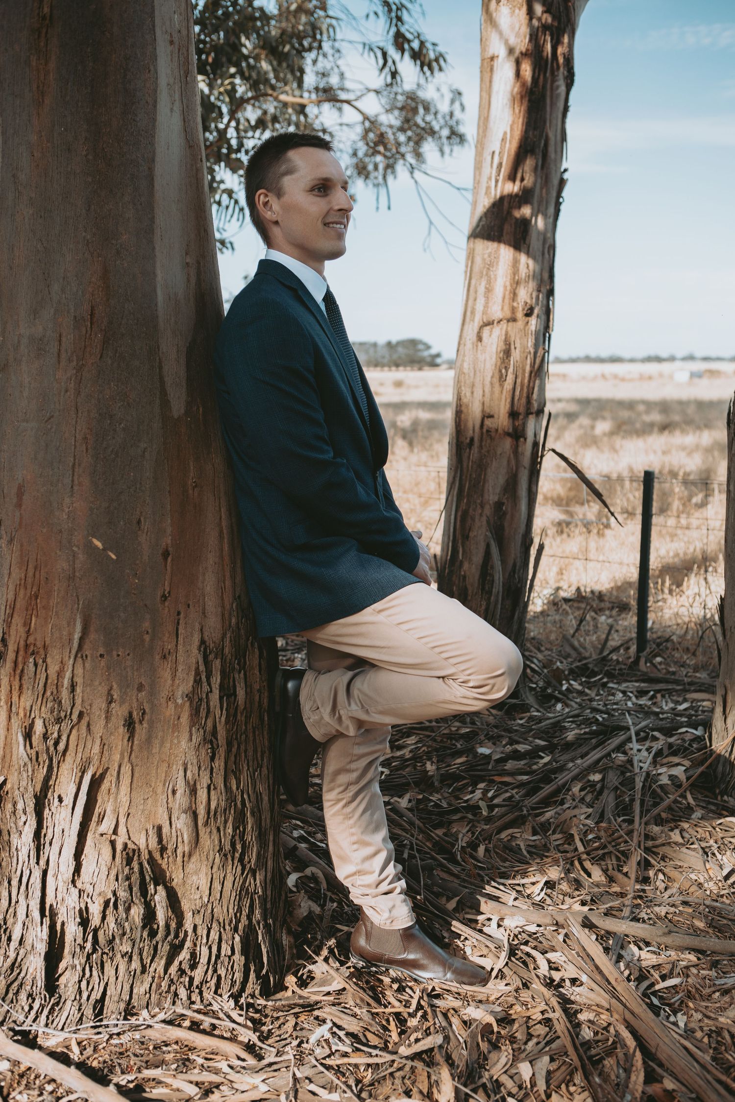Young man in navy blazer and khaki pants leaning against a eucalyptus tree in a rural Australian field.
