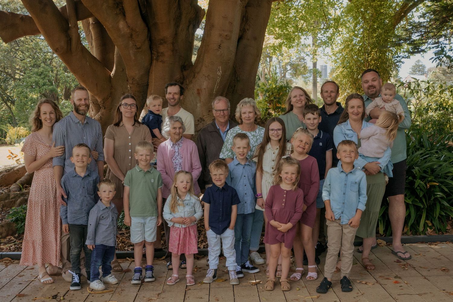 Large multigenerational family portrait gathered outdoors under a sprawling tree on a sunny day at Warrnambool Botanical Gardens.