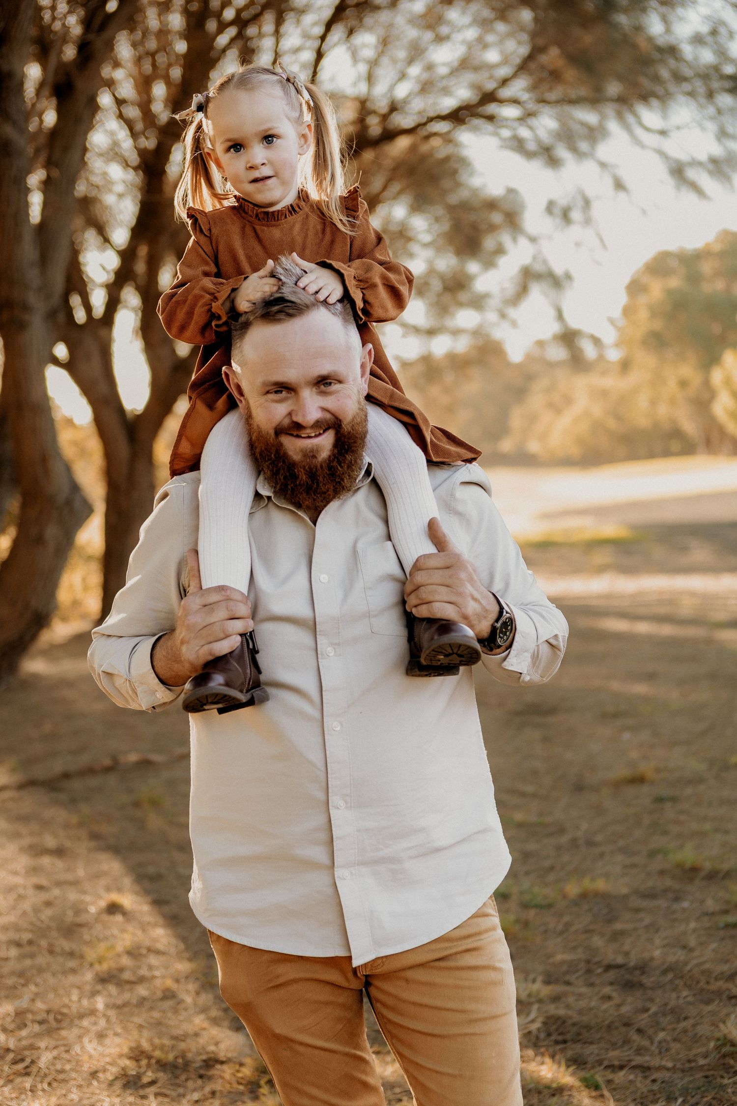 Smiling bearded father carrying his young daughter on his shoulders in golden sunset light.