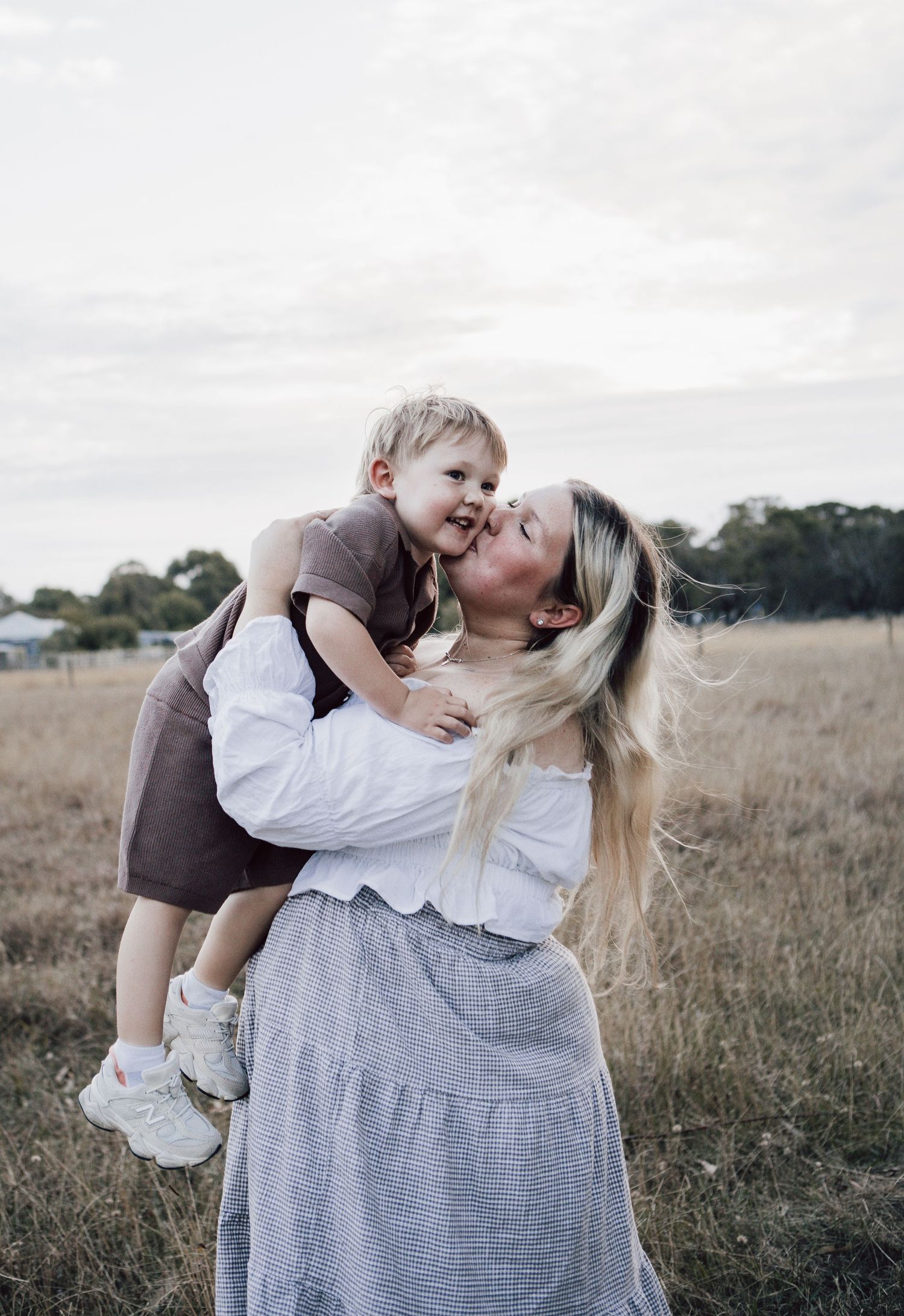 A blonde mother kisses her laughing toddler son while holding him in a golden field.