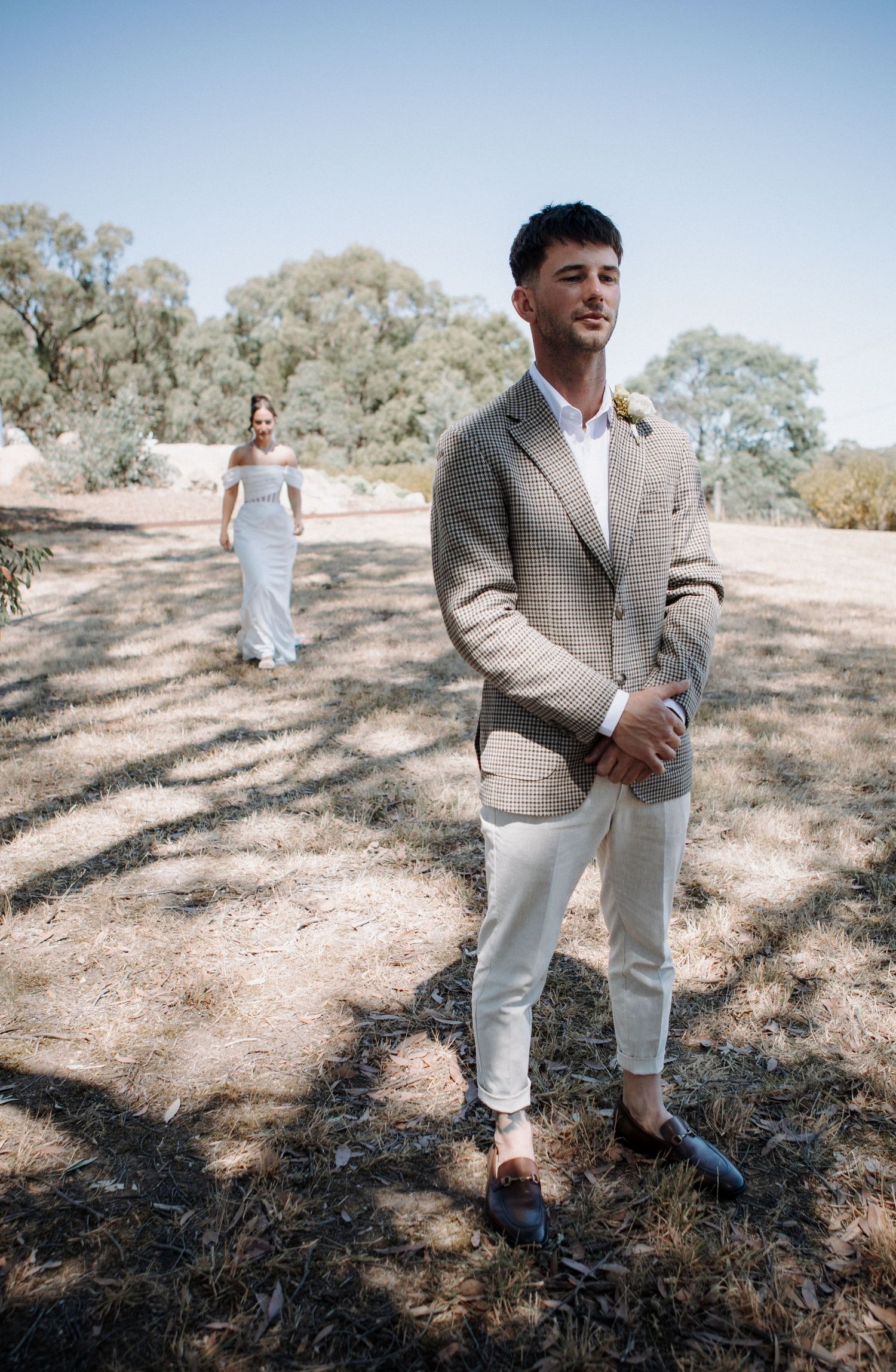 Groom in checkered blazer and white pants stands outdoors as bride in white gown approaches in background.