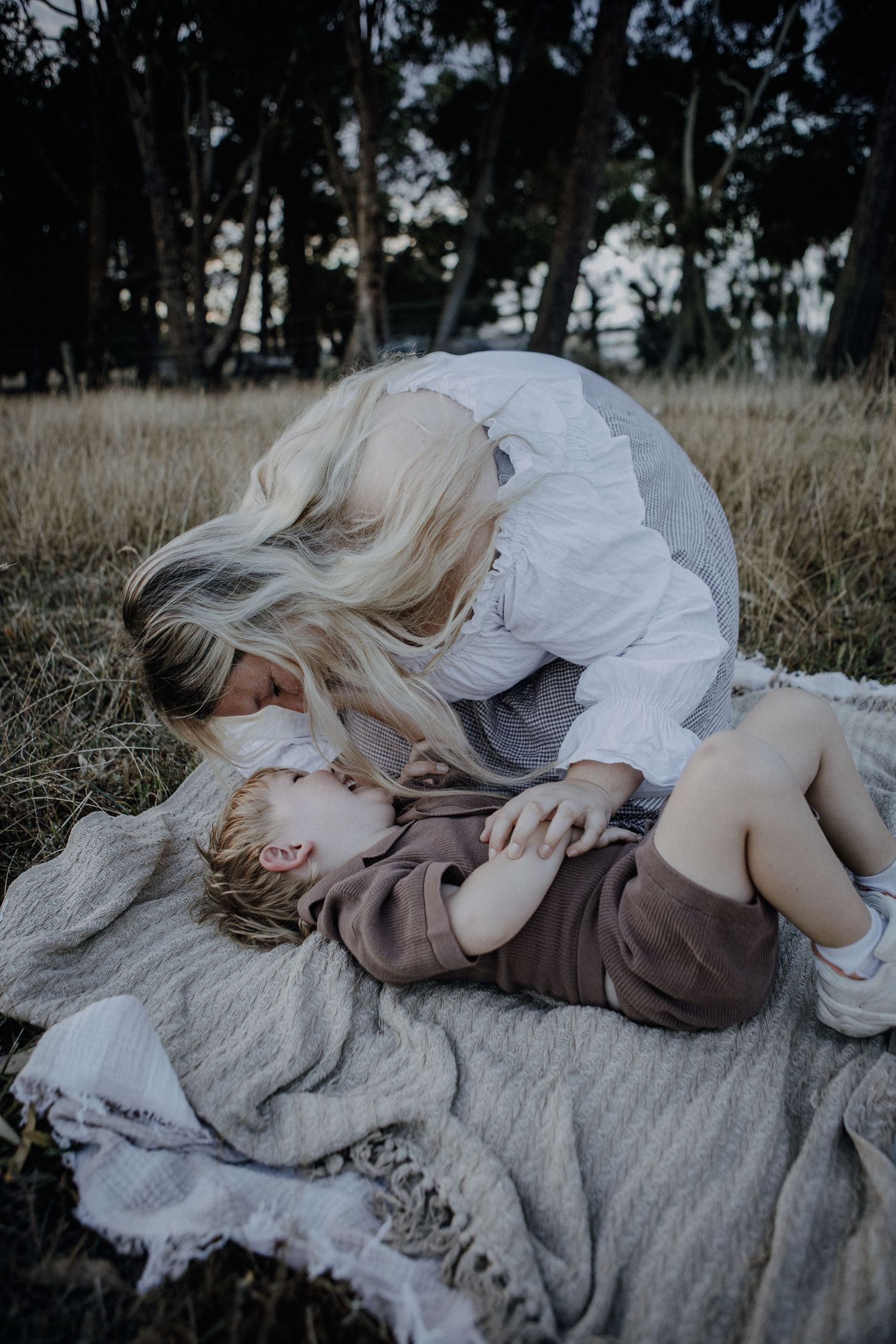 A blonde mother leans down to kiss her toddler lying on a blanket in a grassy field.
