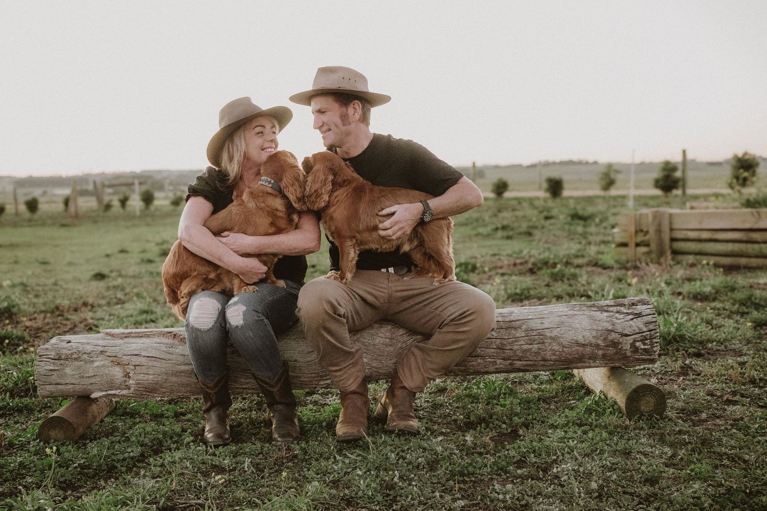 Couple sitting on a log holding brown dogs in a rural farm setting outdoors.