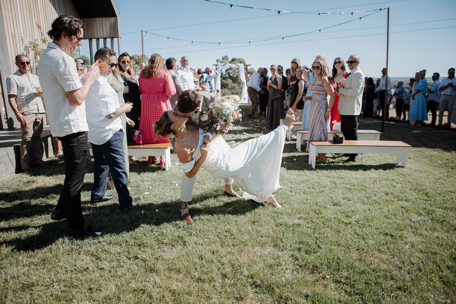 Bride and groom share a romantic dip kiss on grass at outdoor wedding reception with guests watching.