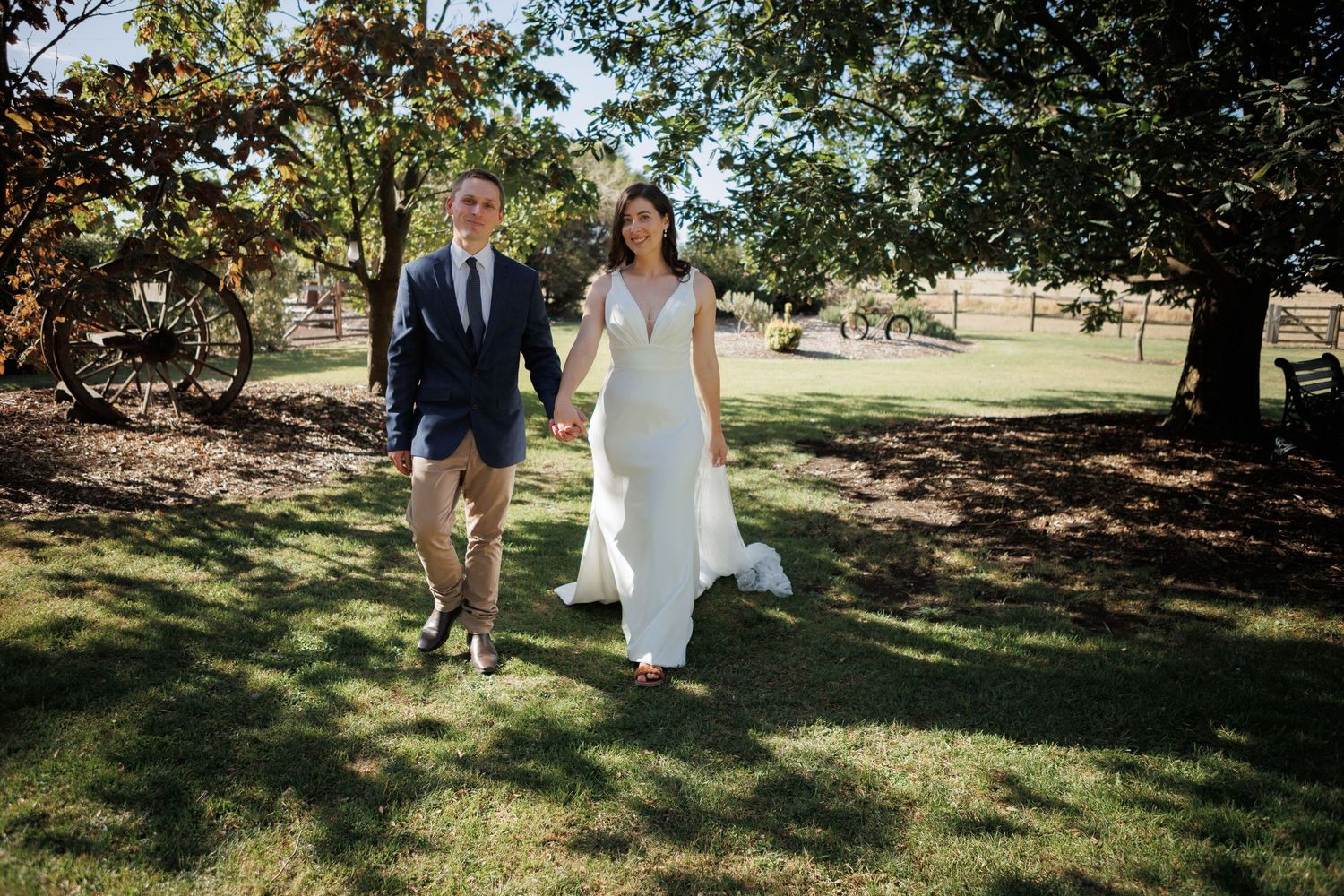 A bride in a white gown and groom in a navy suit walk hand in hand across a green lawn at an outdoor wedding.