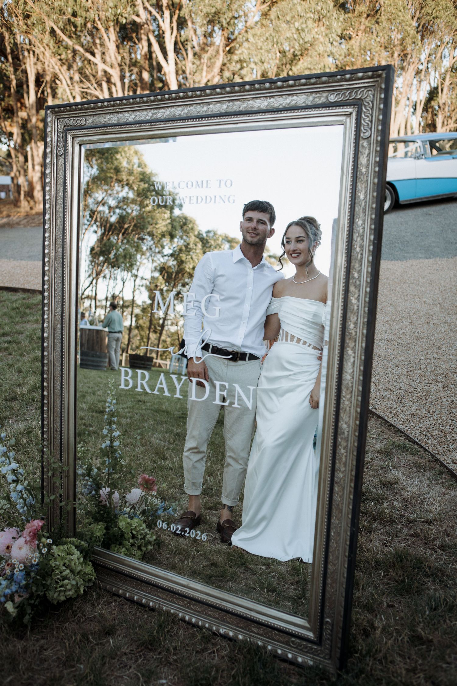 Wedding welcome mirror sign displaying Meg and Brayden with couple reflected in ornate gold frame outdoors.