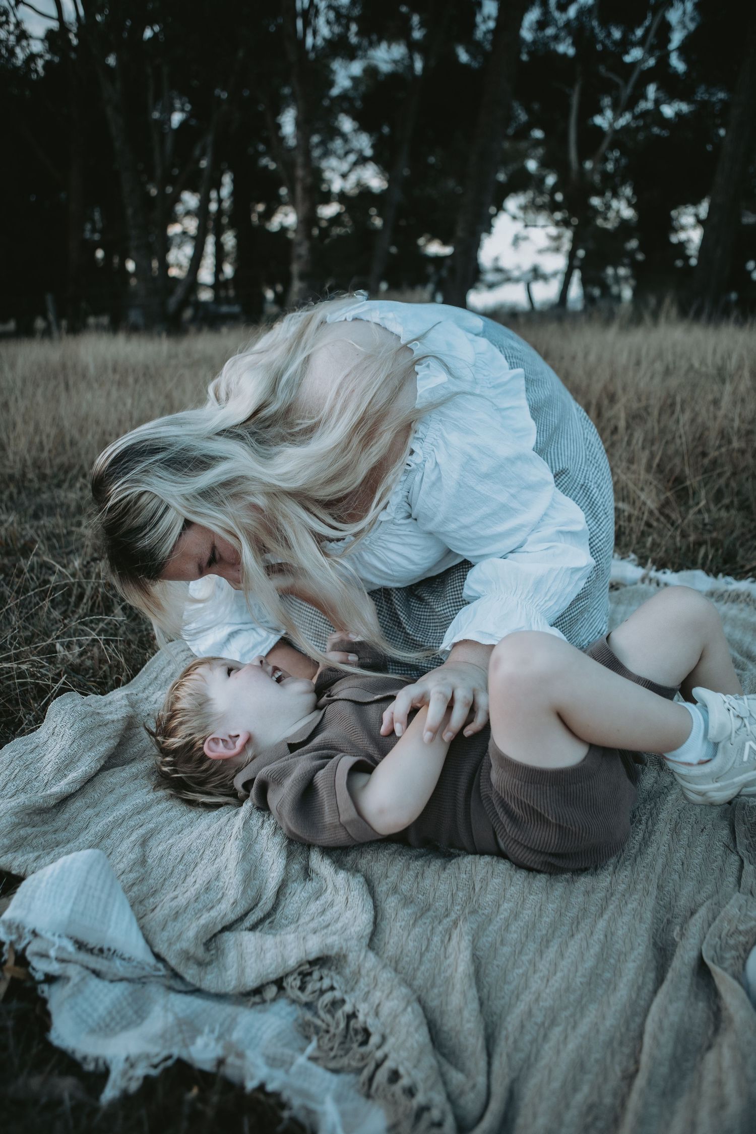 A blonde mother leans over her laughing toddler on a blanket in a grassy field surrounded by trees.