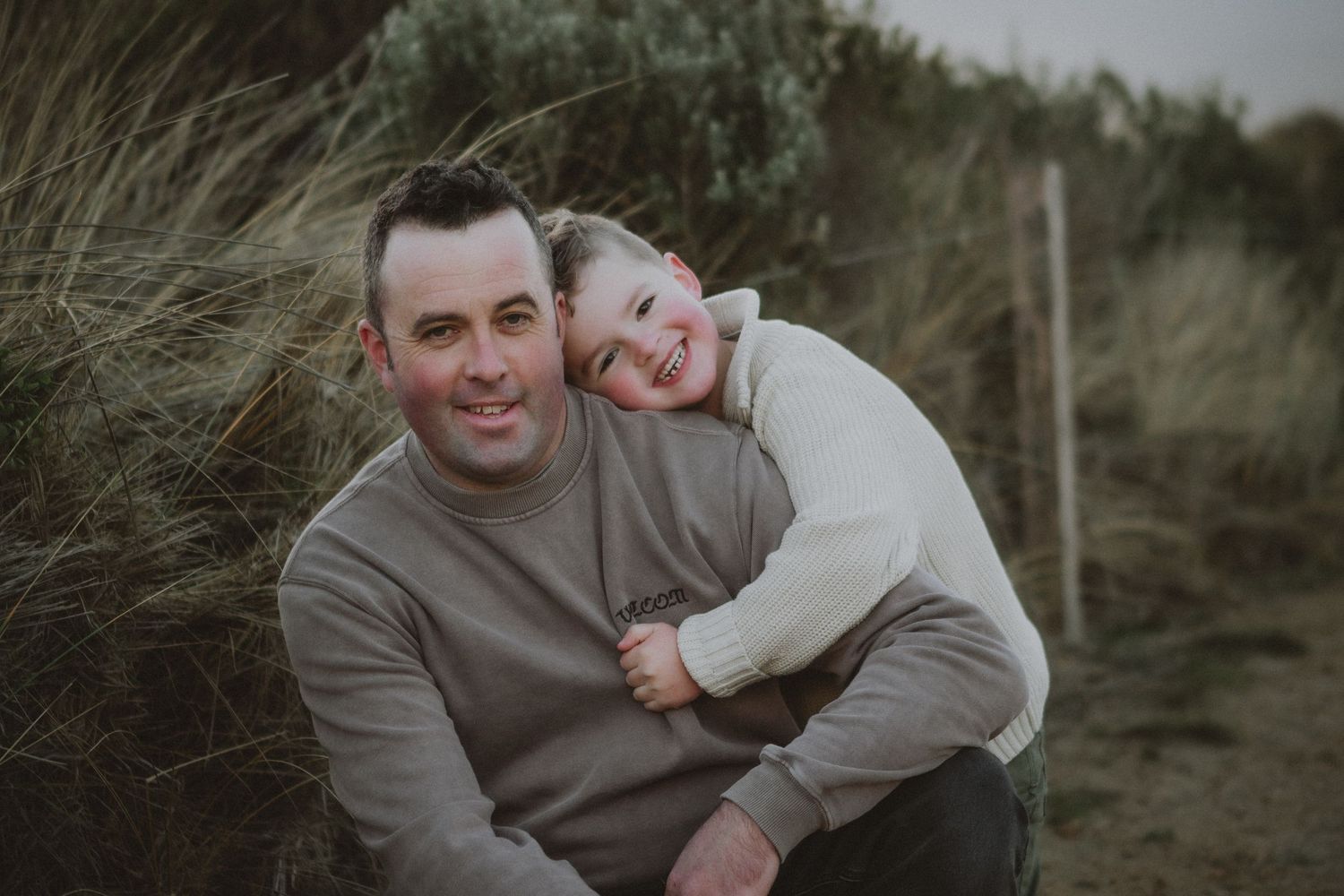 Father and young child sharing a joyful embrace among tall grass dunes in a candid outdoor portrait.