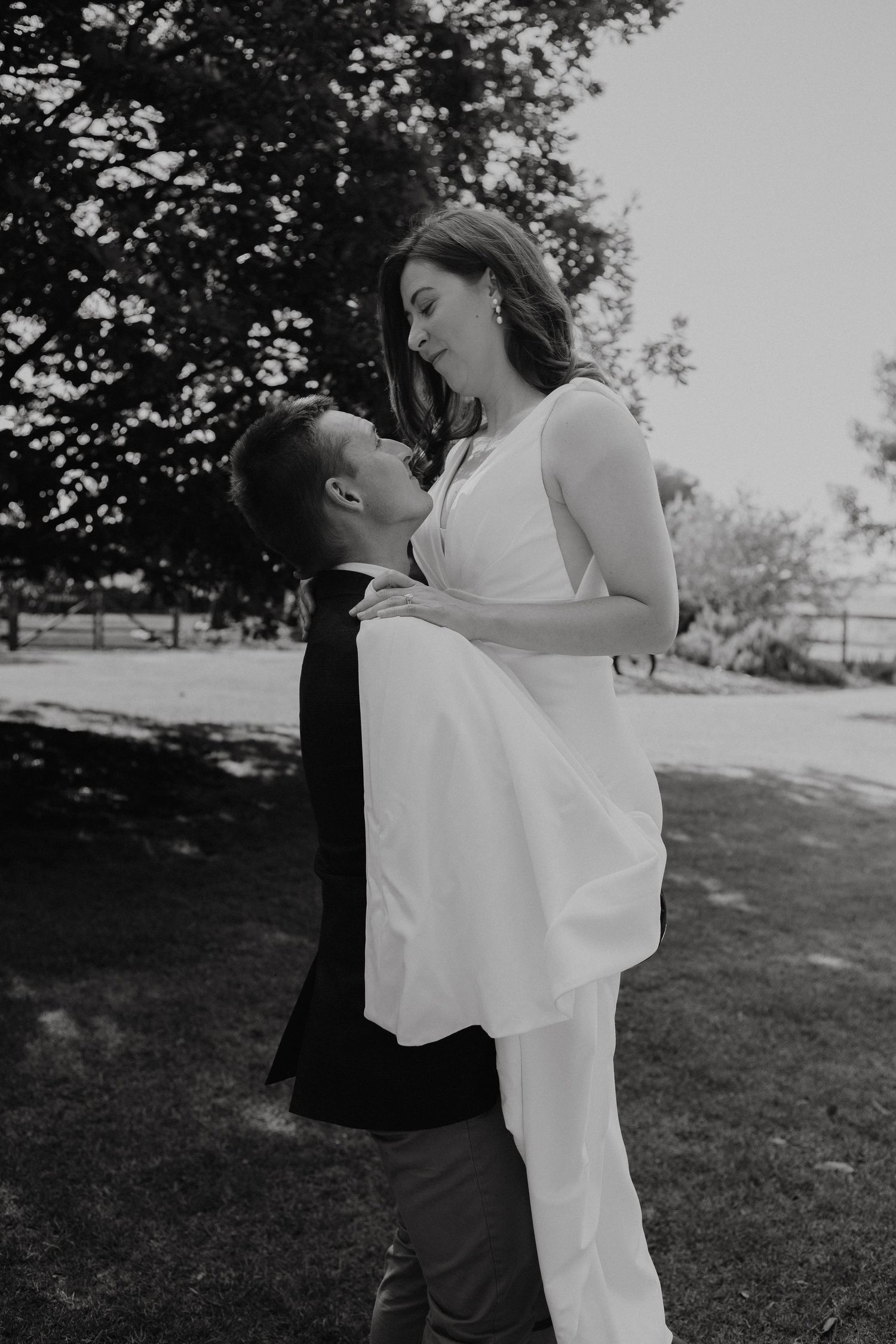 Black and white photo of a groom lifting his bride in a white dress outdoors on their wedding day.