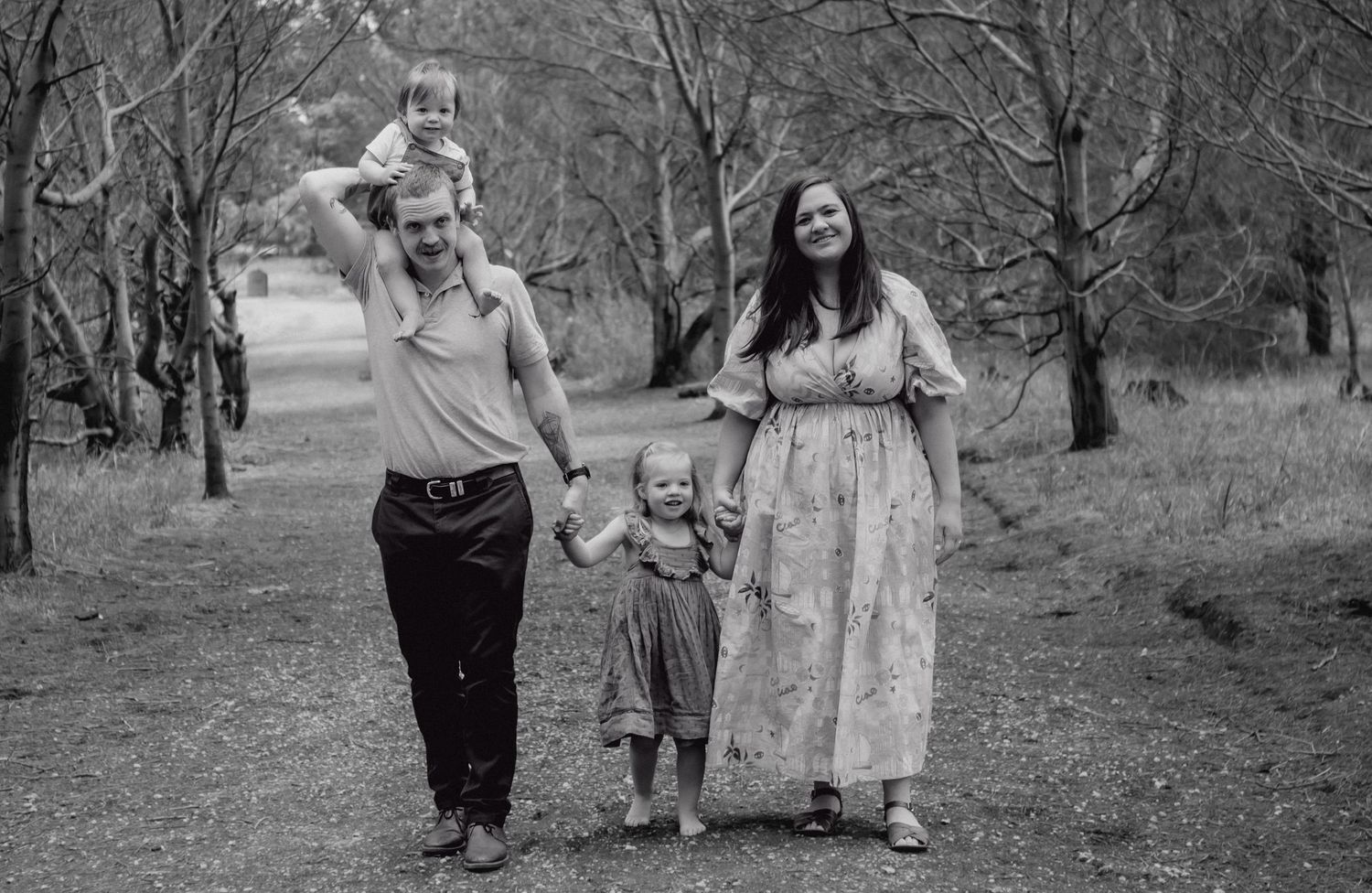 Young family of four walking hand in hand along woodland path in black and white.