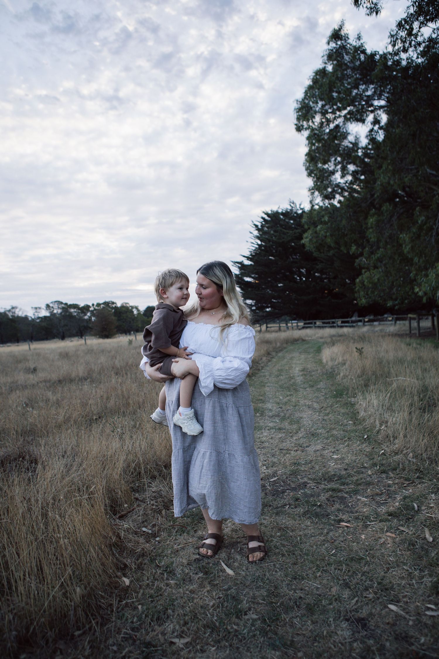 A blonde mother in a white top holds her young toddler son in a golden grass field.