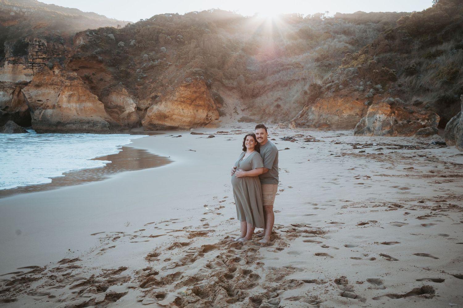 Couple embracing on a sandy beach at golden hour with dramatic cliffs and ocean waves in the background.