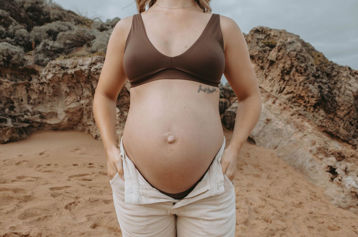 Pregnant woman in brown bikini top and white pants posing on sandy beach with rocky cliffs behind her.