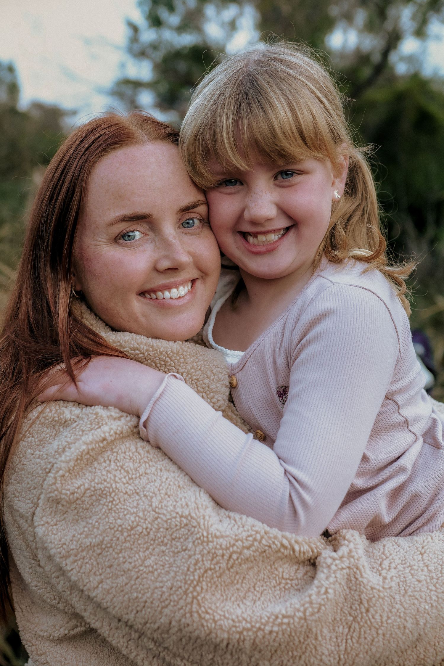Smiling red-haired mother holding her blonde daughter close in a warm outdoor portrait embrace.