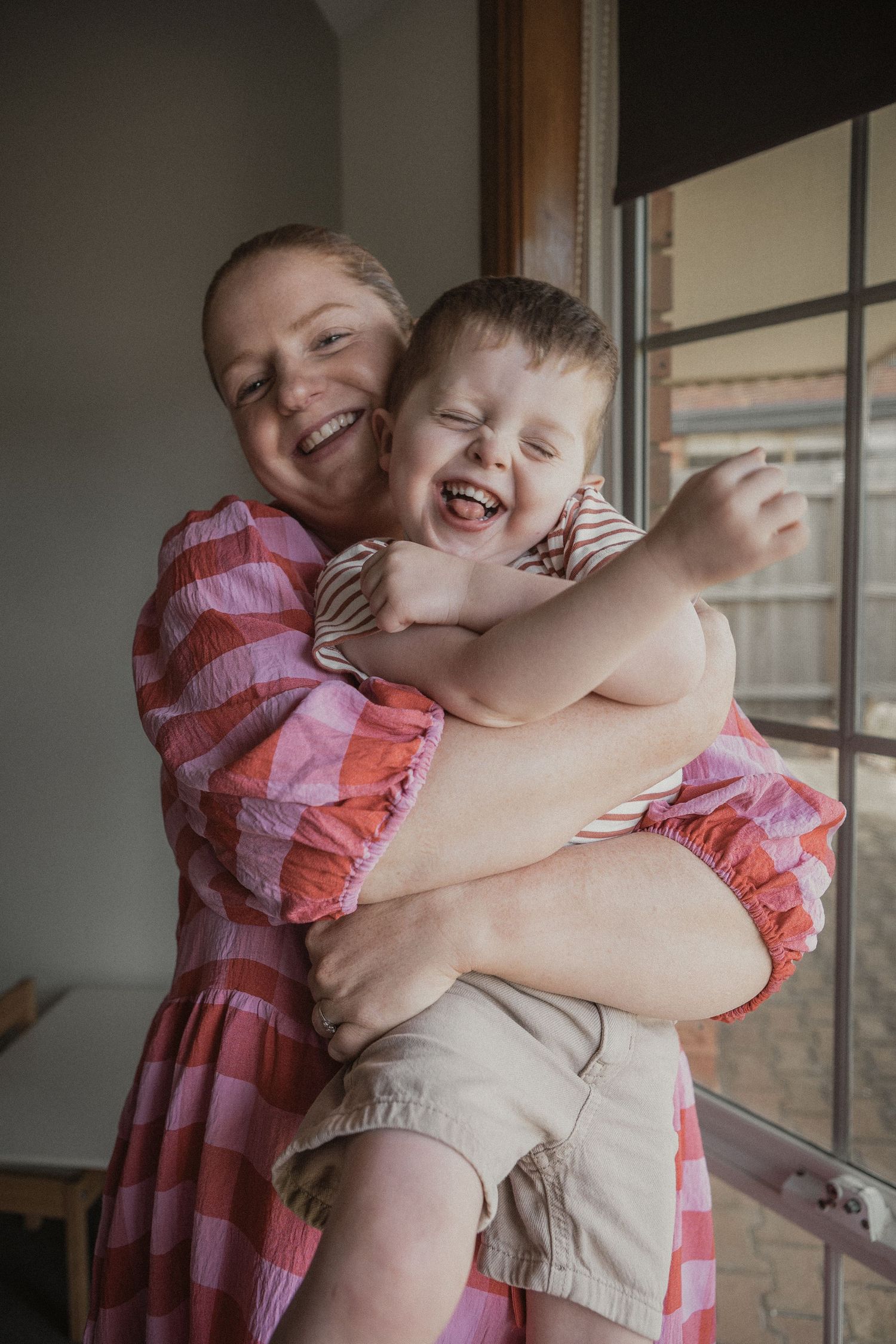 Joyful mother in pink dress hugging laughing young son near window in candid family moment.