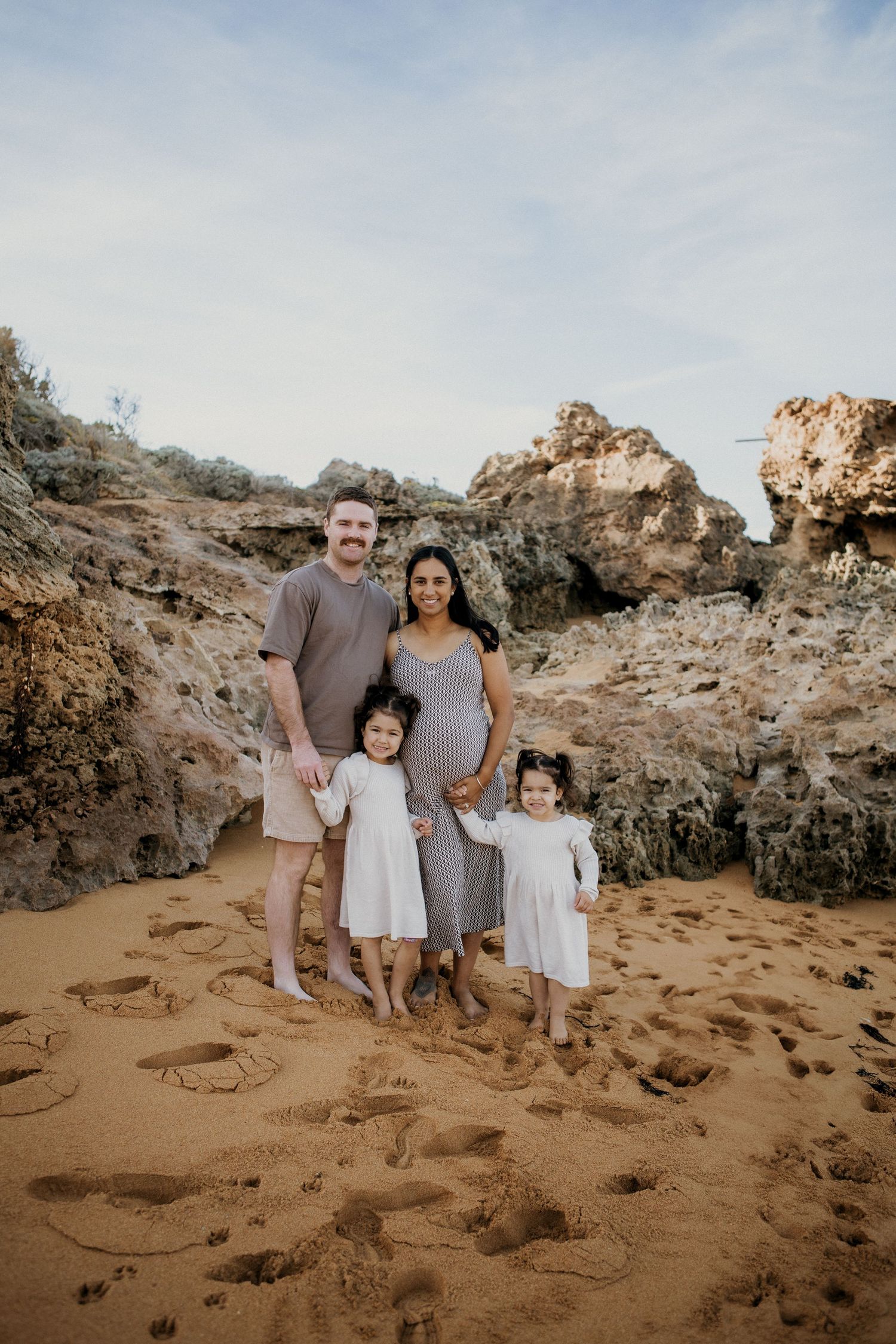 Smiling family of four with expectant mother standing on golden sand beach with rocky backdrop.