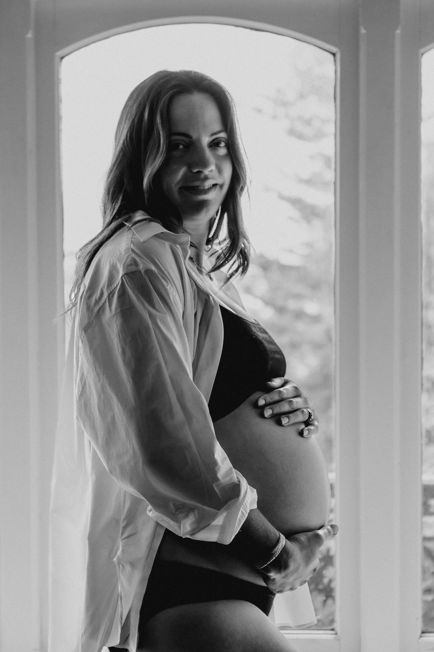 Black and white portrait of a pregnant woman cradling her baby bump by a bright arched window.