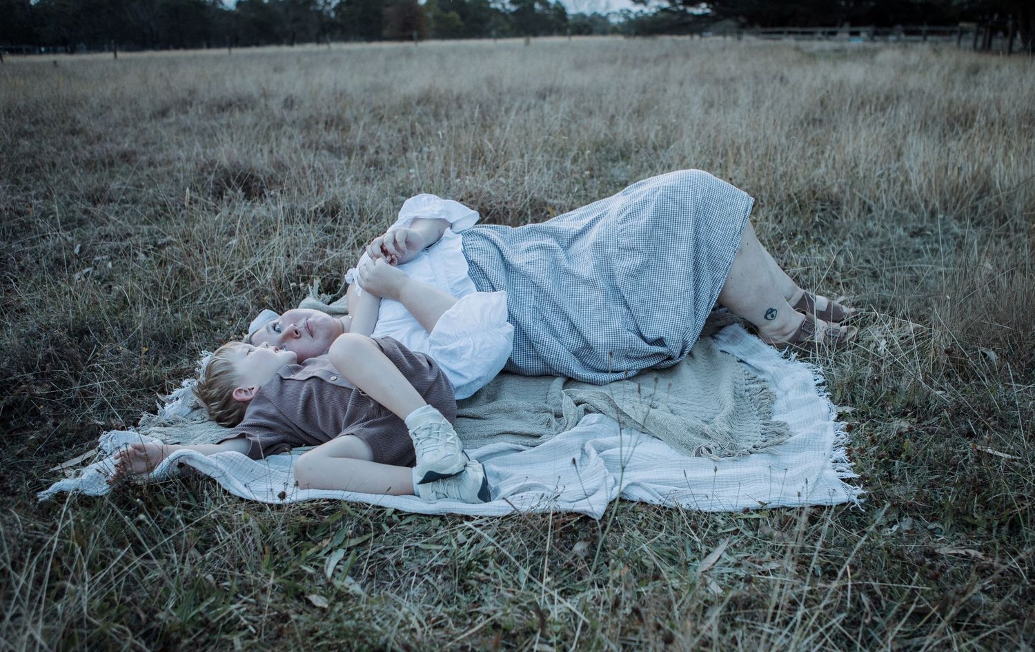 A mother and young child lying together on a blanket in a grassy outdoor field.