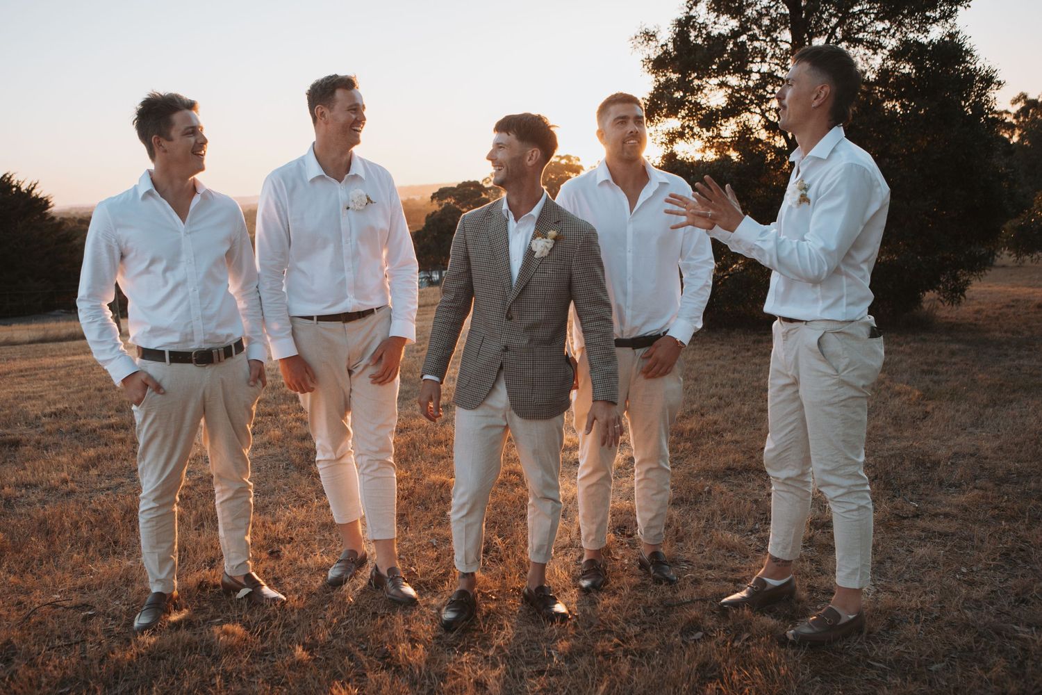 Groom in grey suit stands with groomsmen in white shirts at golden hour outdoor wedding.