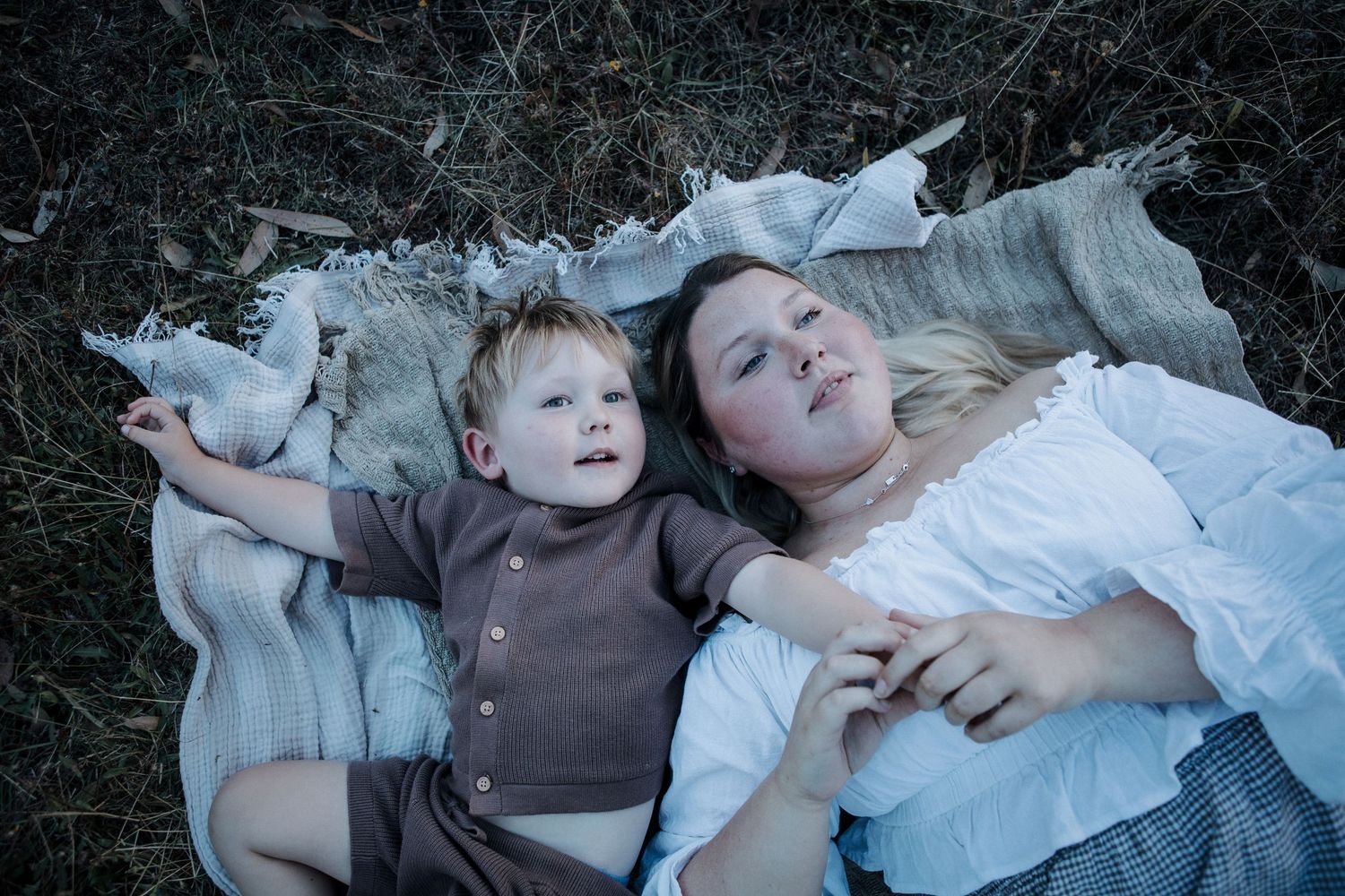 A blonde mother and toddler boy lying on a blanket in a field, relaxing together outdoors.