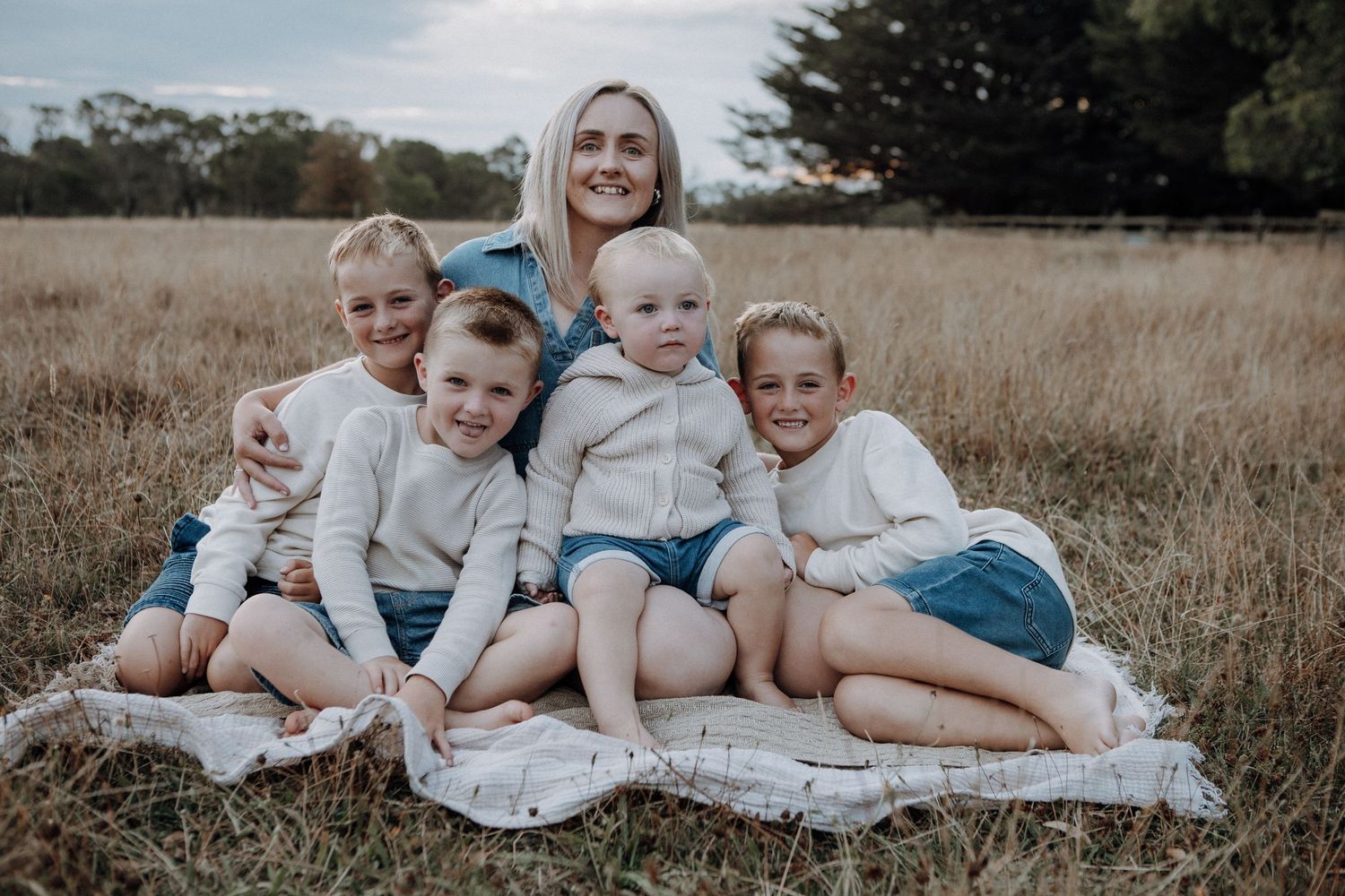 A smiling mother sits on a blanket in a field surrounded by her four young children.
