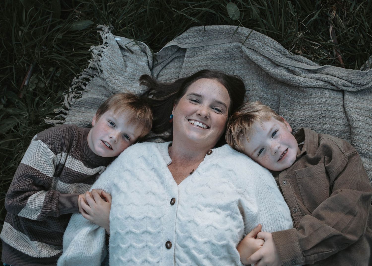 A smiling woman lies on a blanket in grass with two young boys resting their heads on her shoulders.