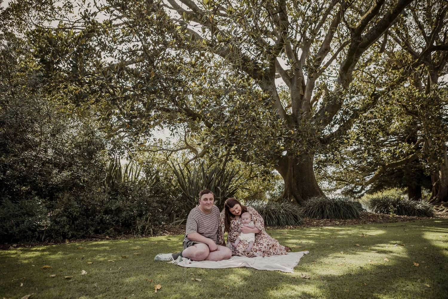 Young couple sitting on blanket with toddler under large oak tree in lush green at Warrnambool Botanical Gardens.