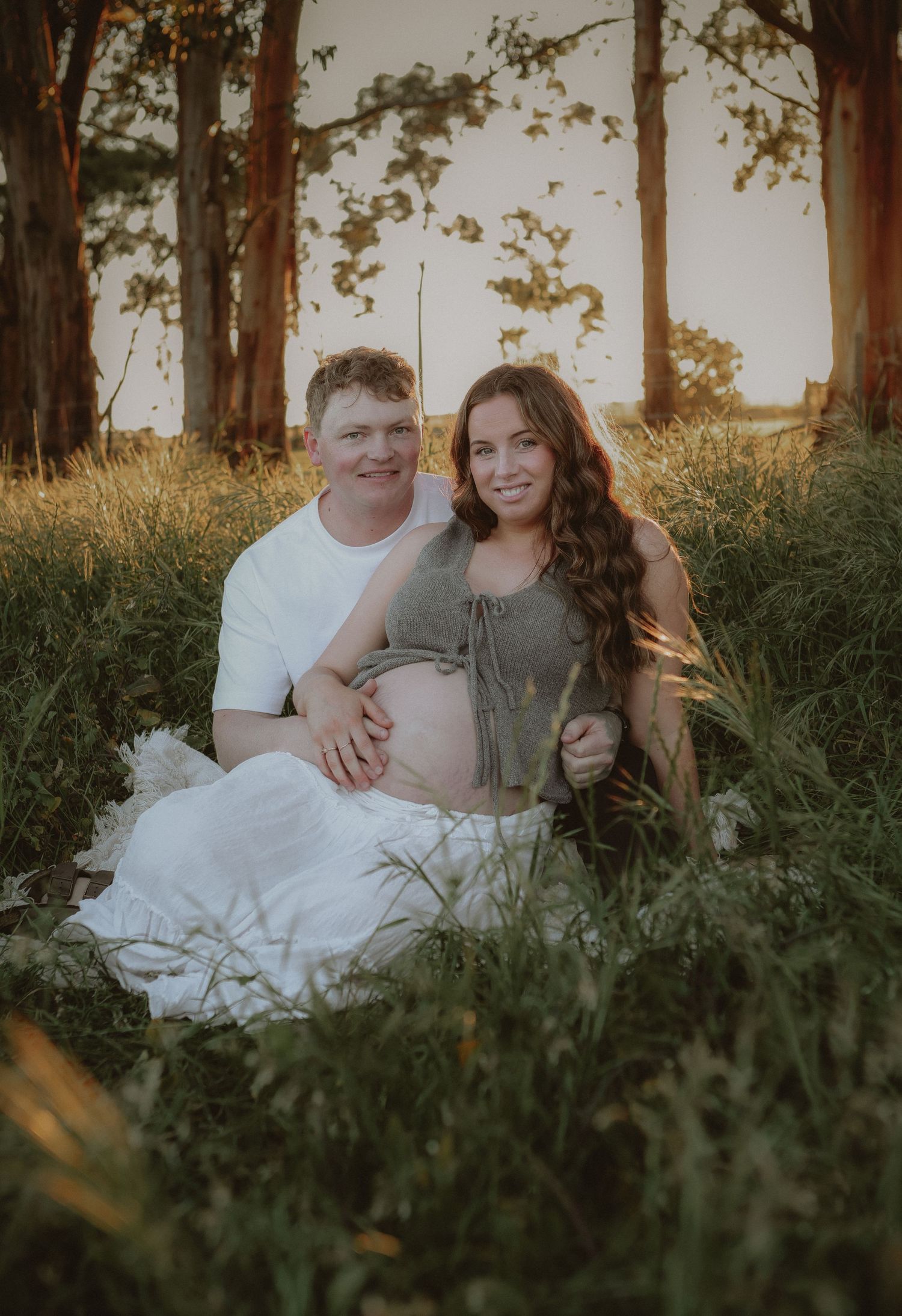 Expecting couple sitting in golden sunset meadow, woman showing baby bump in maternity photo session.