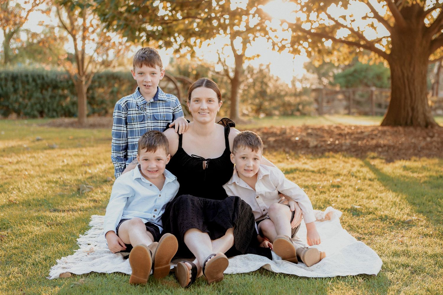 Mother sitting on grass with three young boys in outdoor park setting during golden hour.