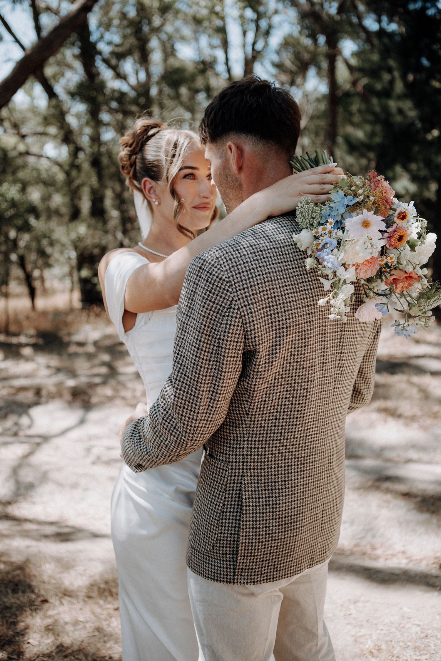 Bride holding a colorful wildflower bouquet embraces her groom in a checkered blazer among pine trees.