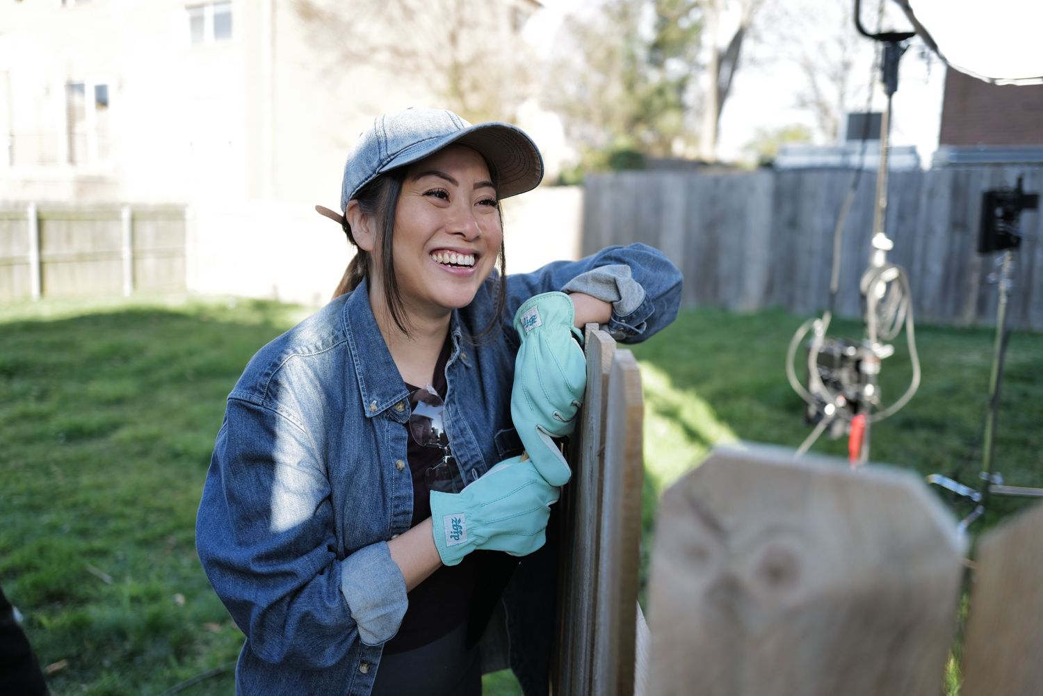 Woman in denim jacket and cap smiling while working on a fence in a backyard garden.