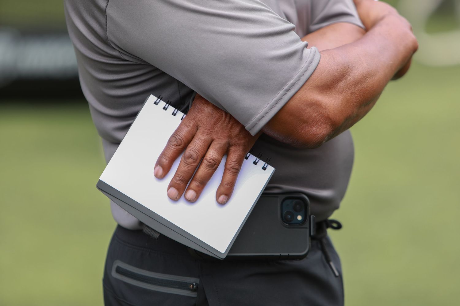Person holding a small notebook and phone against their torso on a sports field.