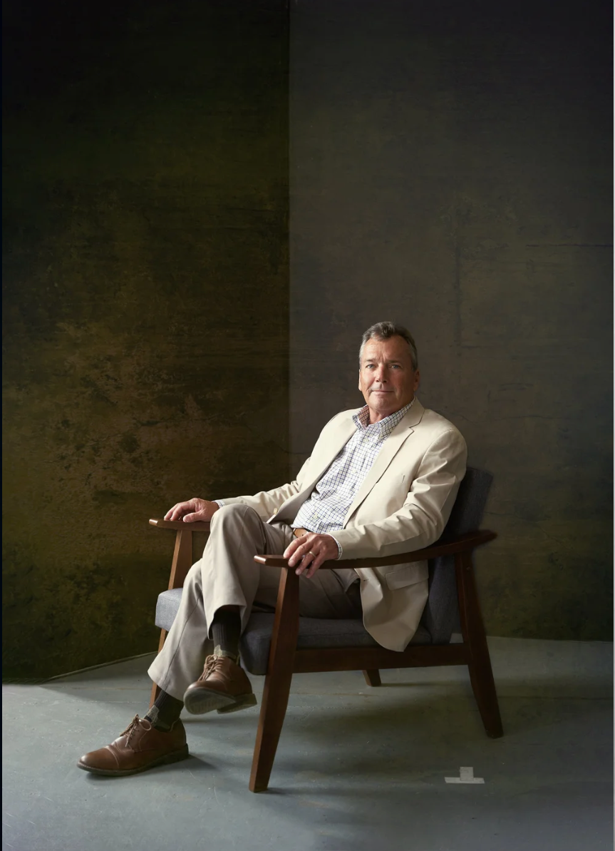 A person in a white suit reclines across multiple positions on a wooden bench against a dark textured wall.