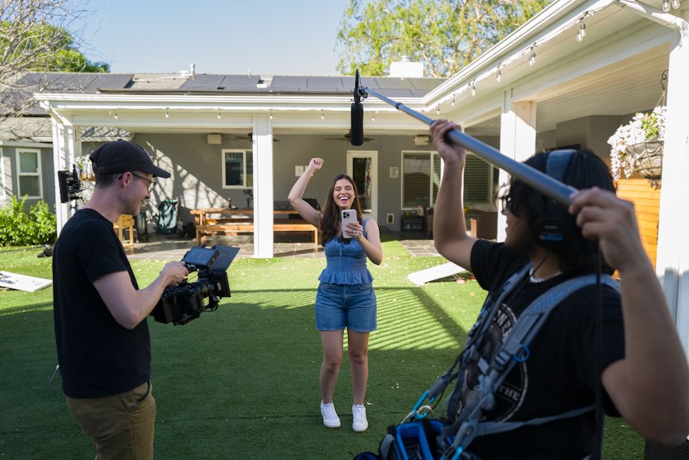 A woman in denim overalls poses excitedly for a video crew filming in a sunny residential backyard.