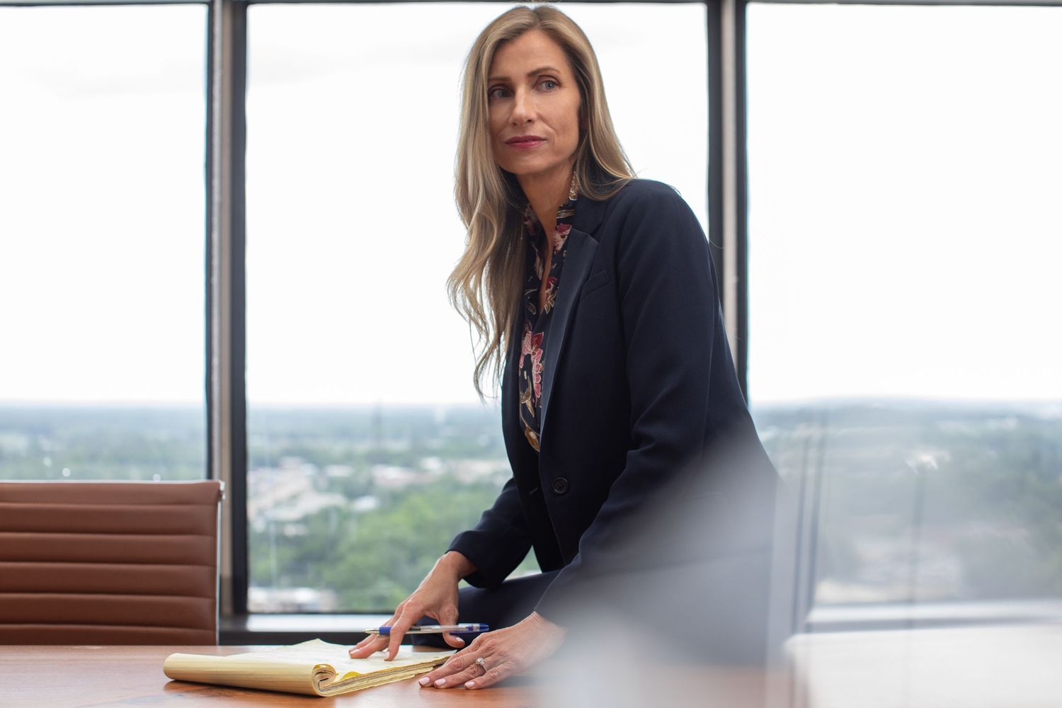 Professional in a dark blazer sits at a conference room table with large windows overlooking a city view.