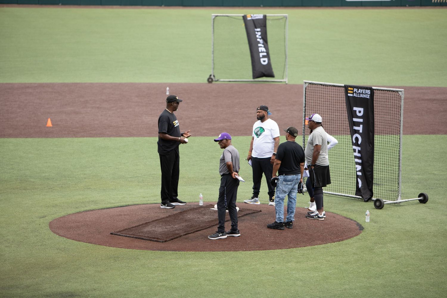 Baseball training session with coaches and players gathered around home plate on a green field.