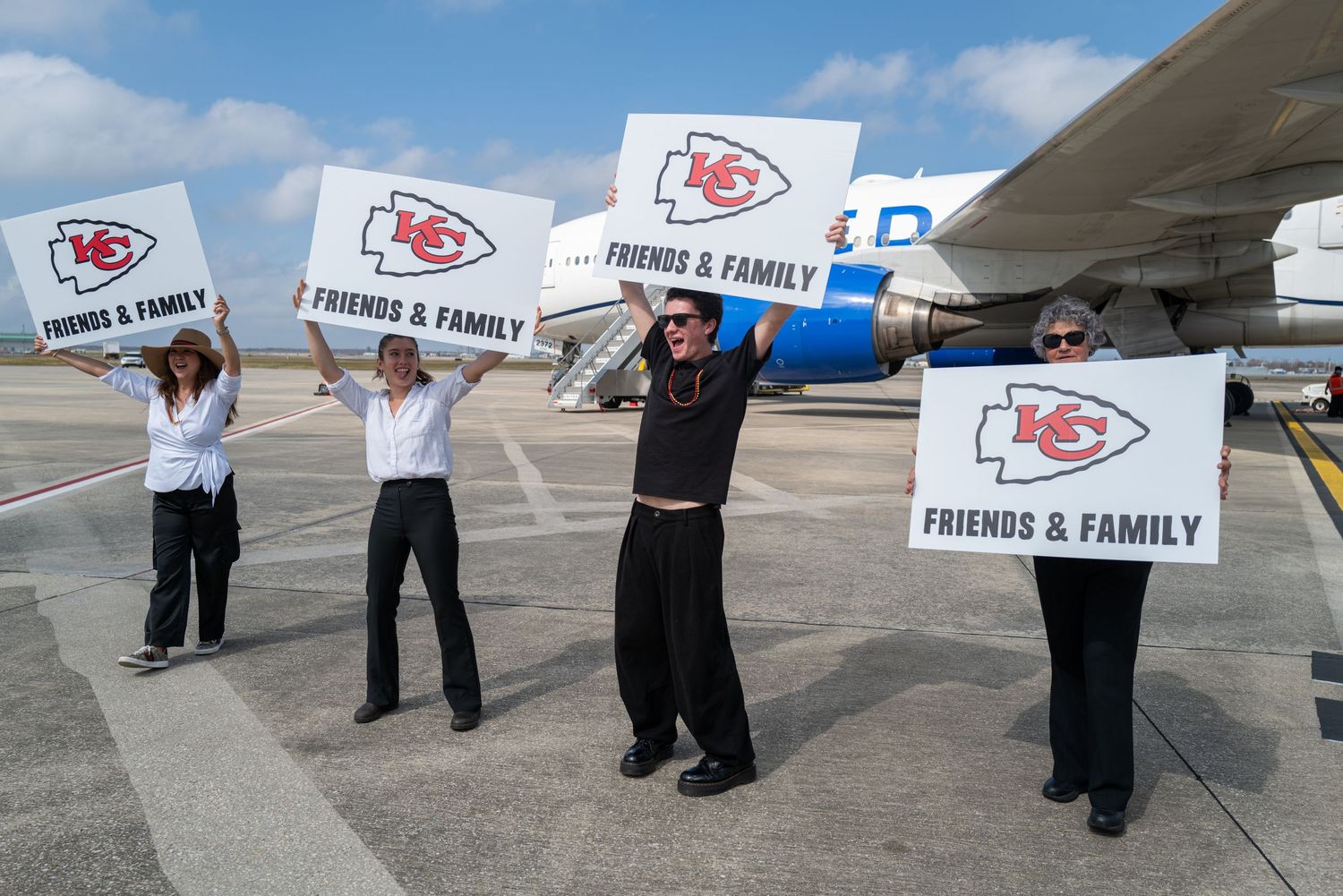 Staff members hold up 'Friends & Family' signs with Chiefs logos on an airport tarmac.