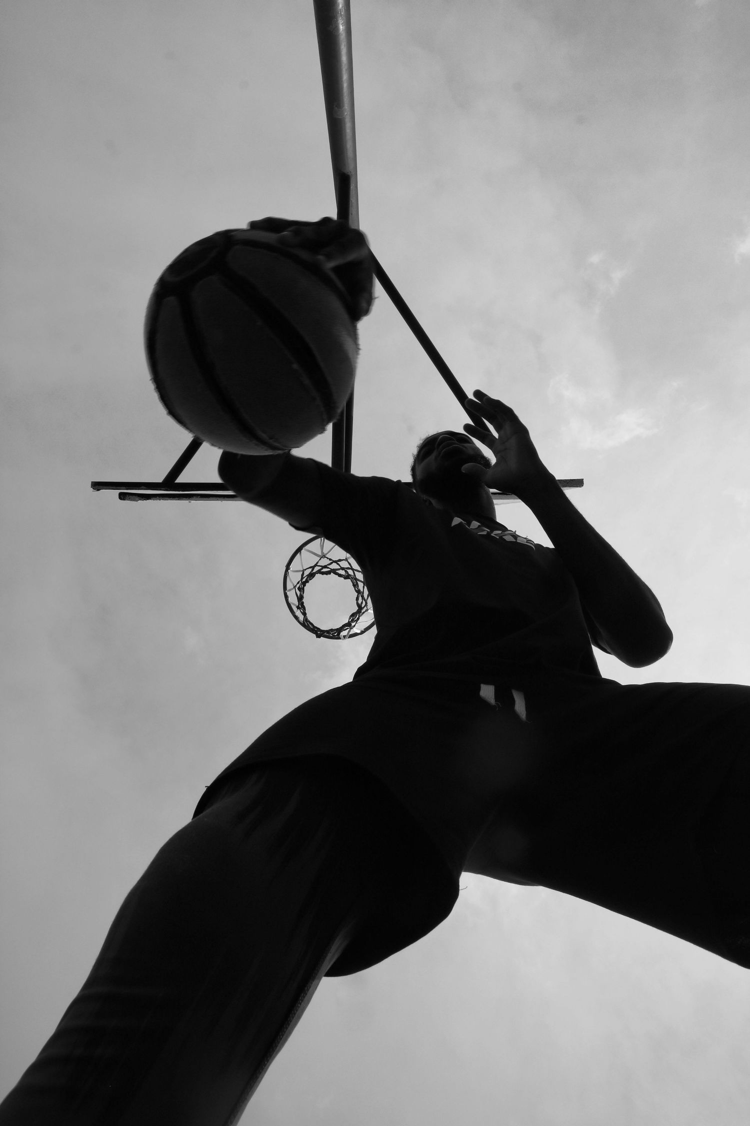 Black and white silhouette of basketball player dunking against cloudy sky.