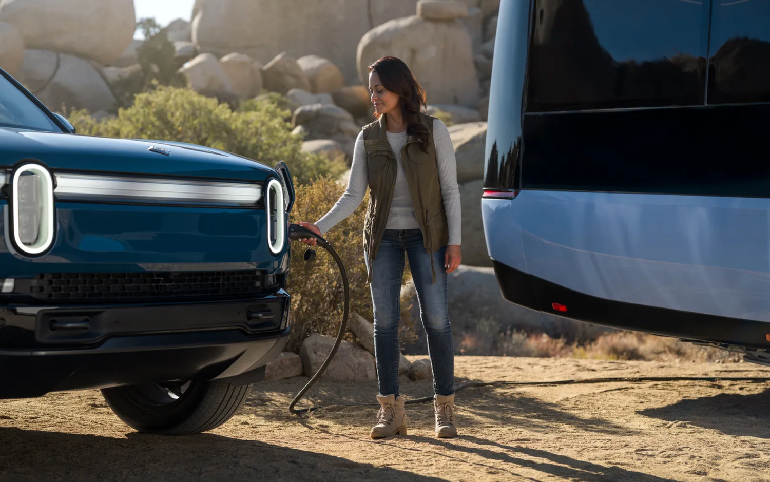 Woman charging a Rivian electric vehicle in a desert landscape next to a silver trailer.