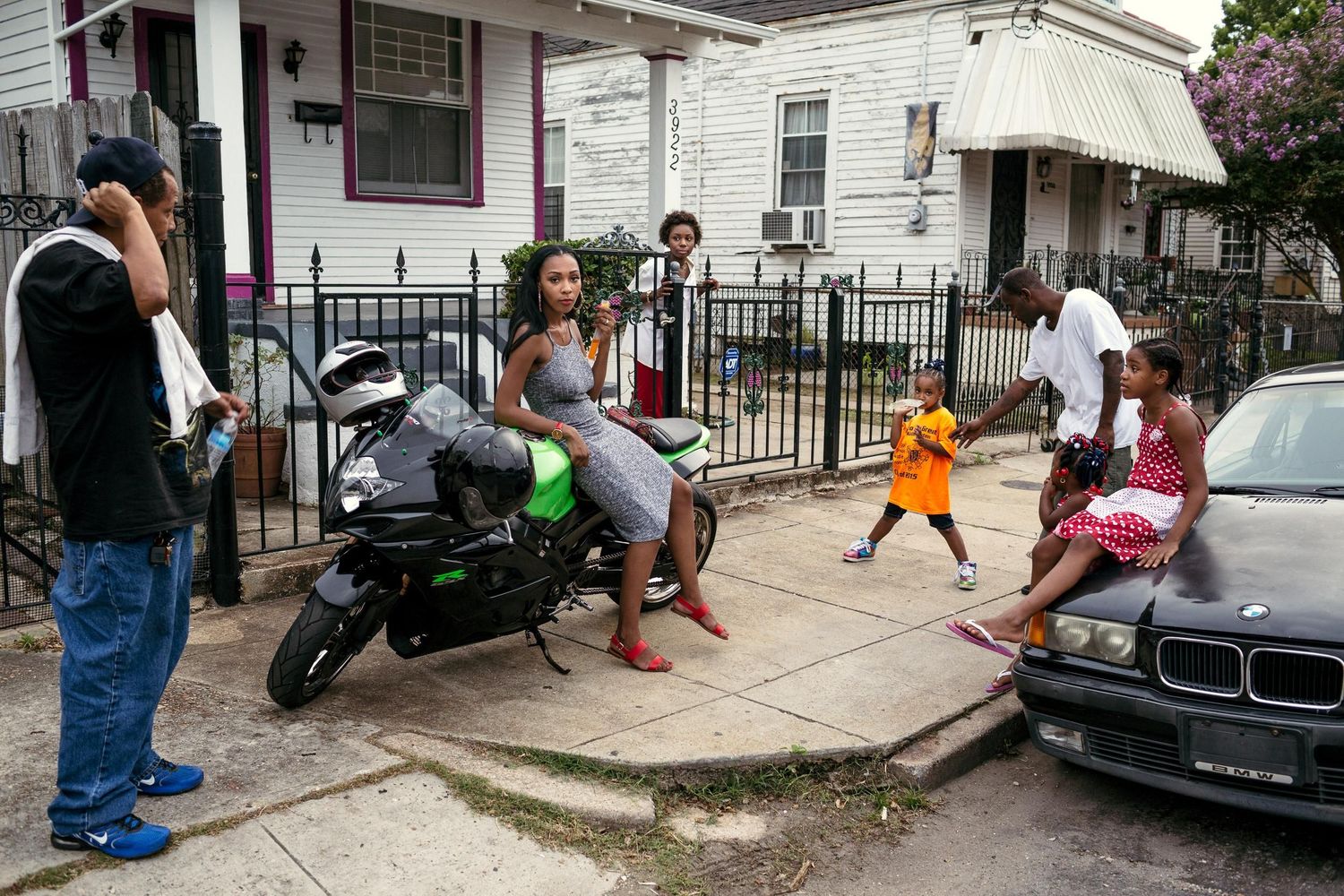 Children play and jump on a residential sidewalk while adults watch from nearby porches and steps.