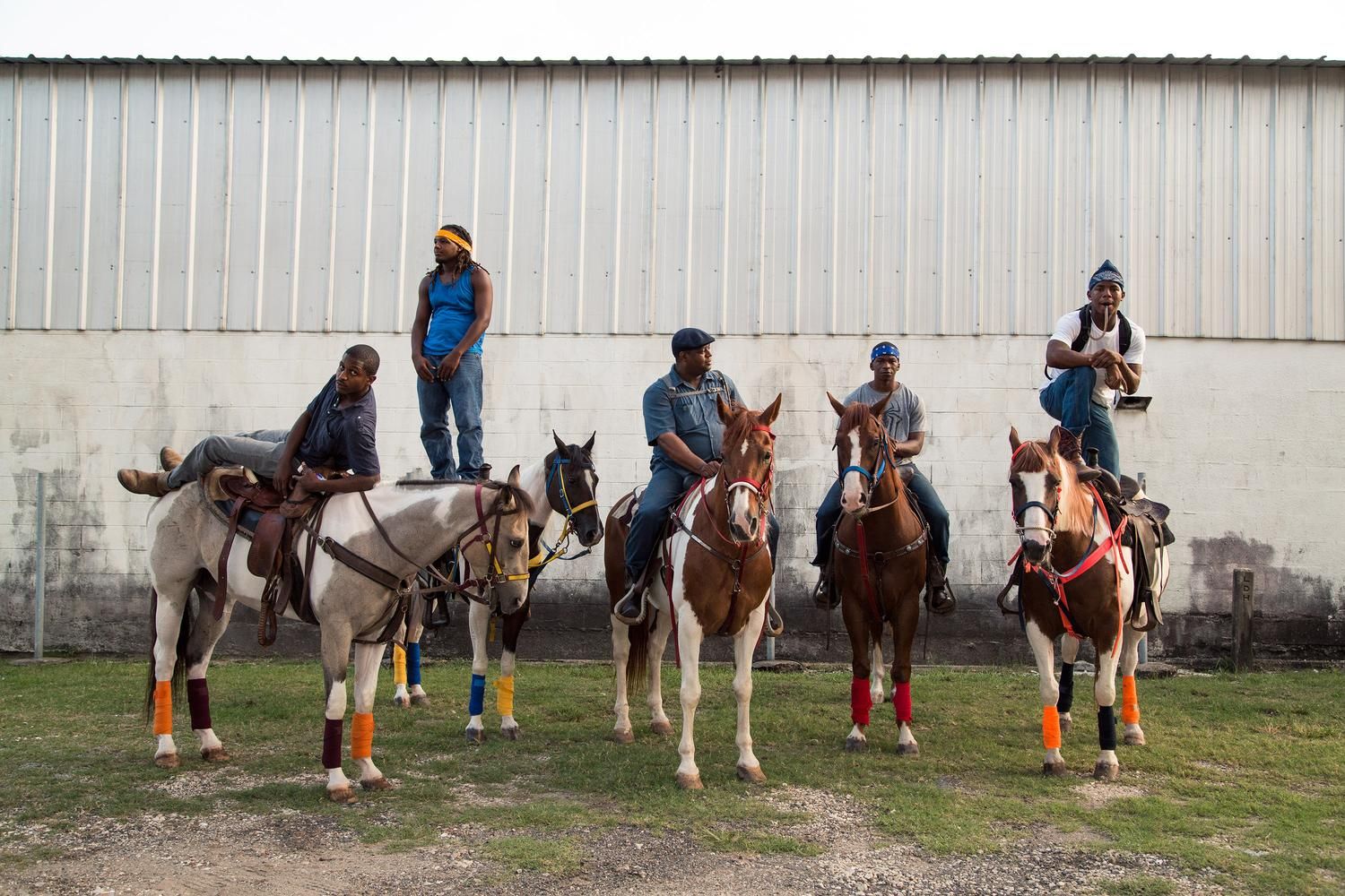 A series of equestrian riders mount their horses in a line against a white building wall.