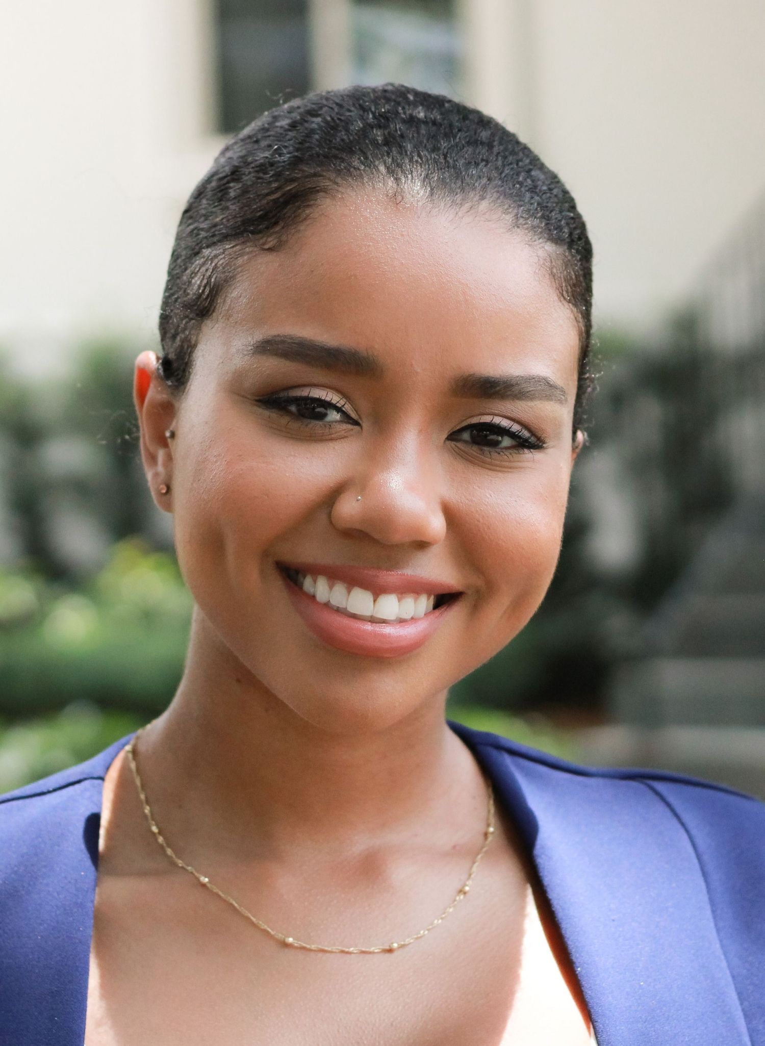 Young woman with short hair smiling outdoors, wearing a blue blazer and gold necklace.