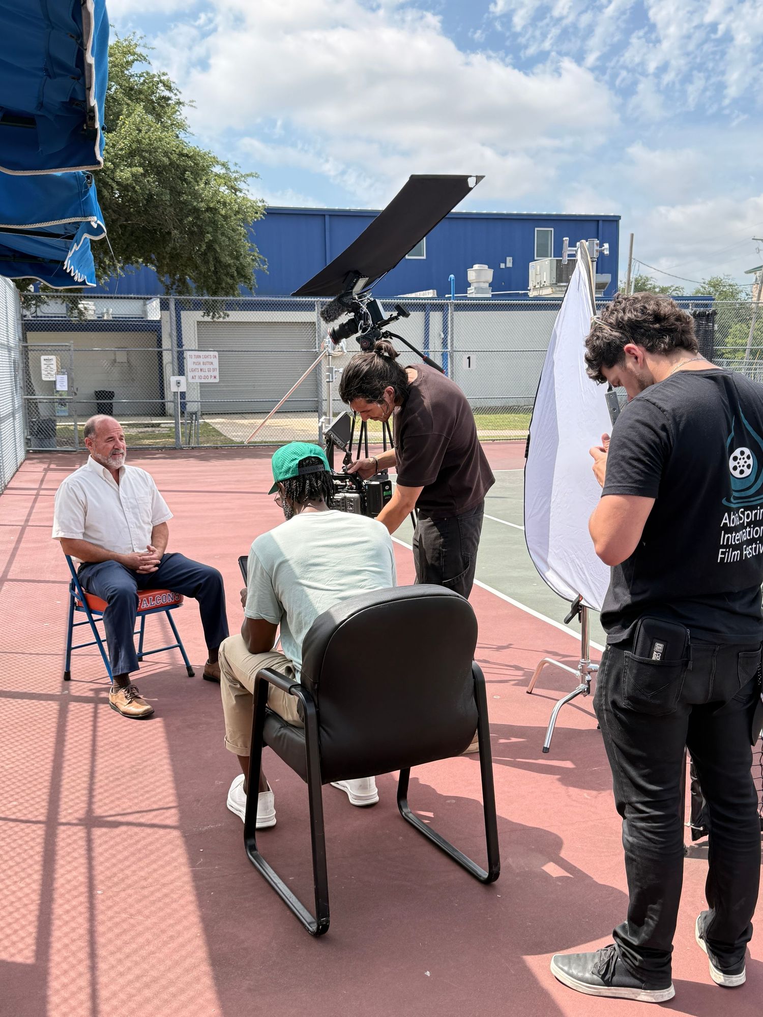 Film crew conducting an outdoor interview on a basketball court with camera equipment and reflector.