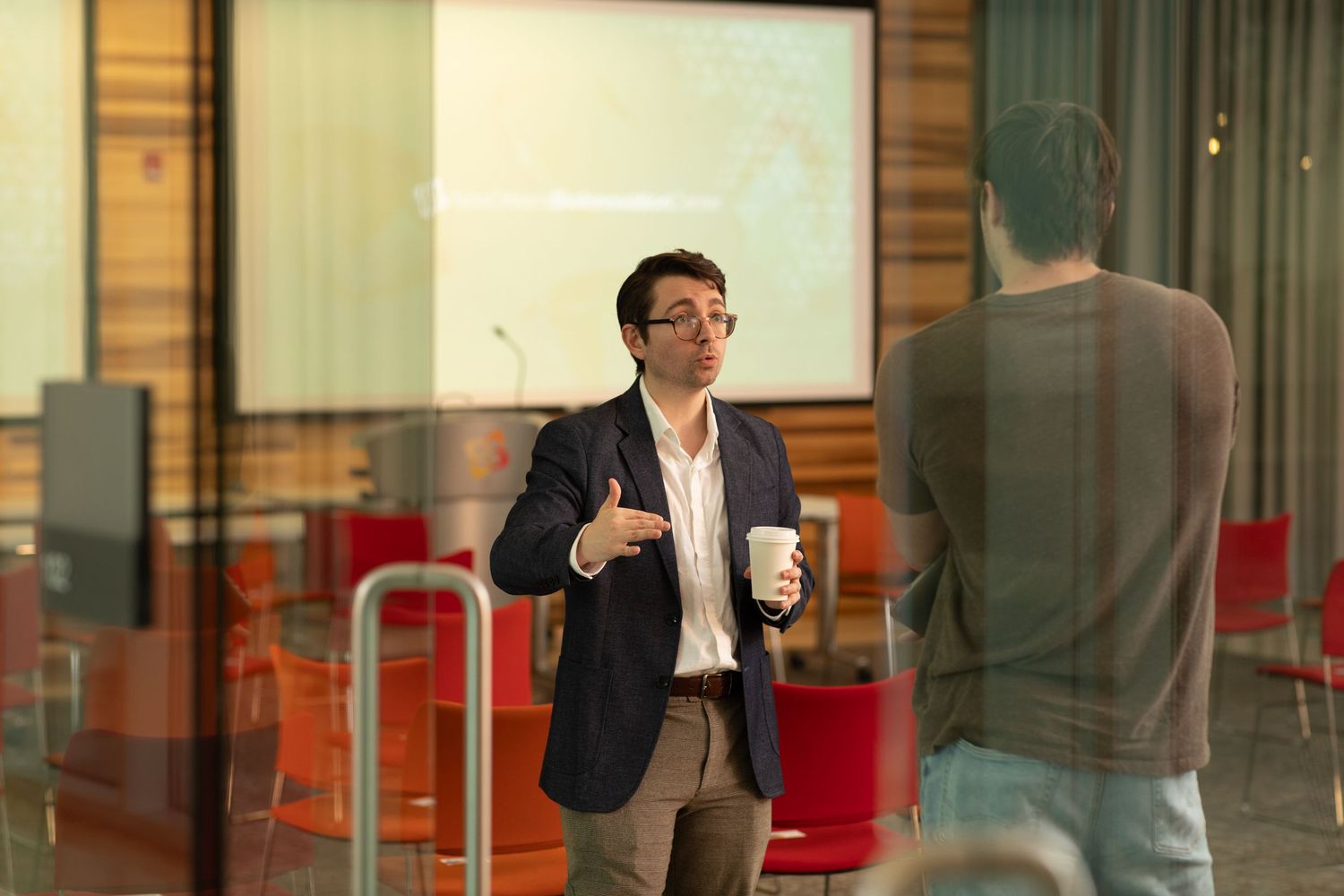 Business professionals having a casual conversation in a modern office hallway with glass walls and red accents.