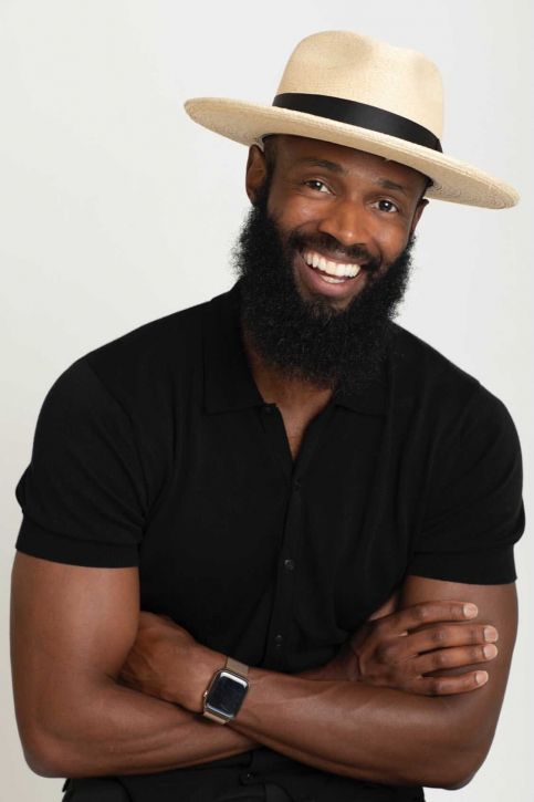 Smiling Black man with long beard wearing straw hat and black polo shirt, arms crossed, studio portrait.